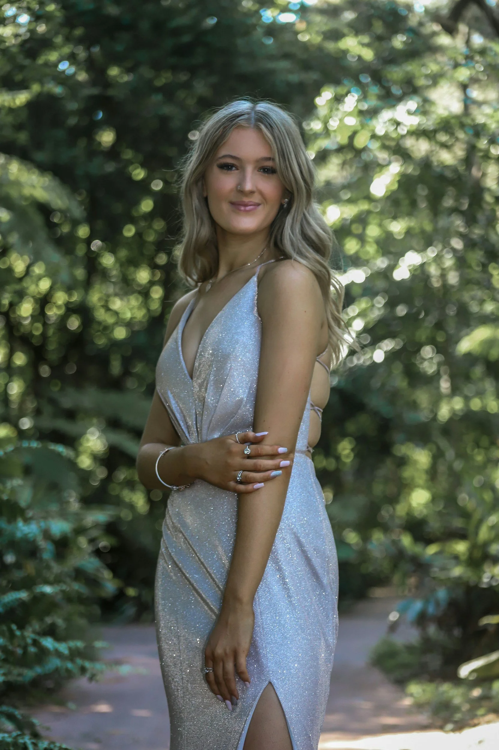 A young woman with long blonde hair, wearing a shiny silver dress with a slit, standing outdoors among green trees, smiling and looking at the camera.