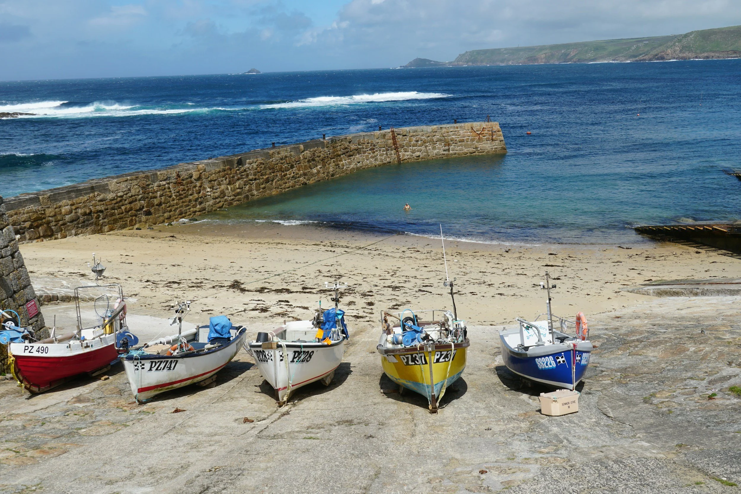 Small boats lined up on a beach near a stone wall with the sea and green cliffs in the background.