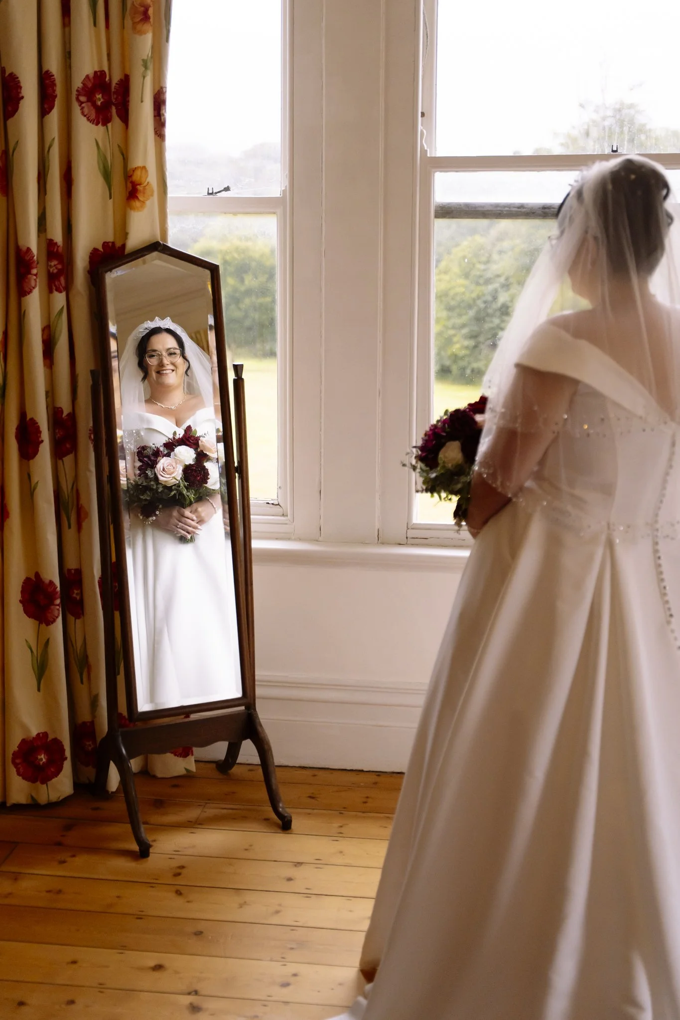 A bride with glasses holding a bouquet of roses and peonies, looking at her reflection in a mirror. She wears a wedding dress with off-shoulder sleeves and a veil, standing in a room with floral curtains and a large window overlooking greenery.