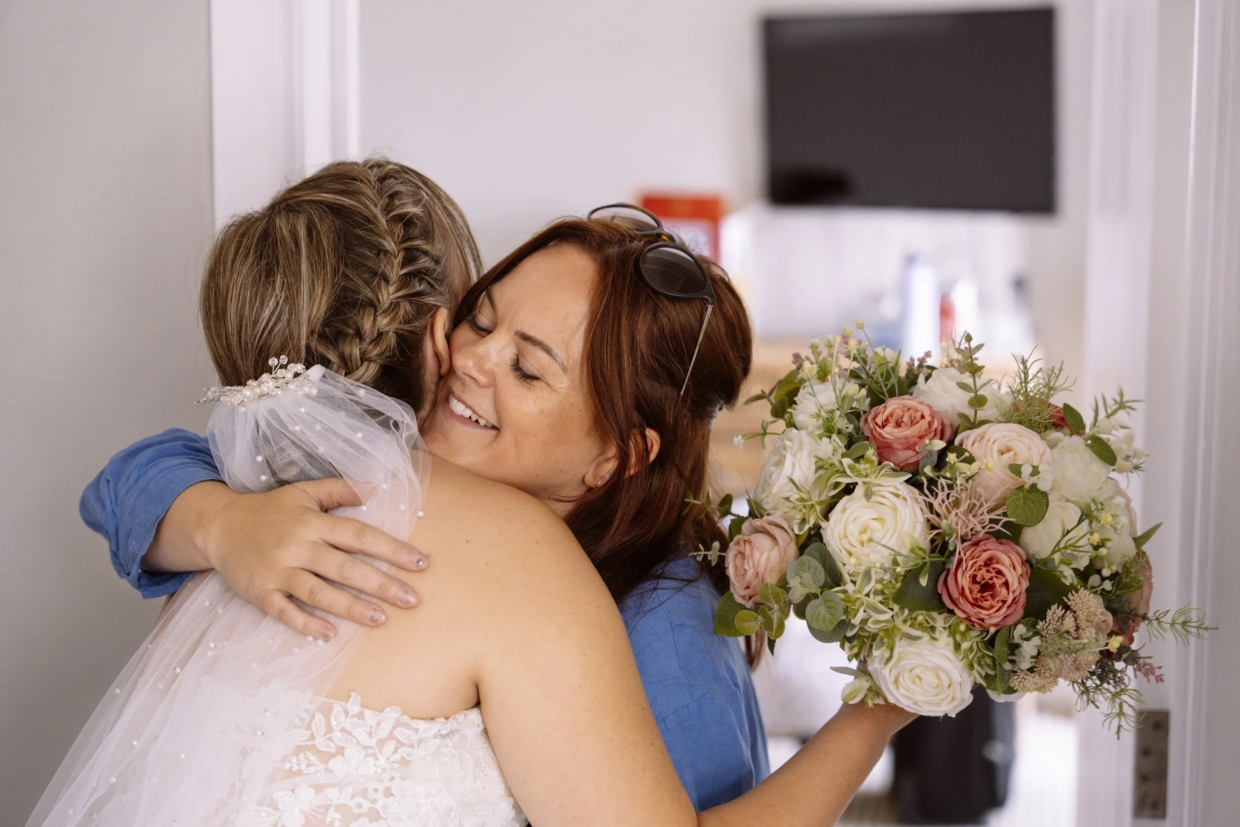 A woman in a blue dress and sunglasses on her head hugging a young girl dressed in a white dress with a pearl headband, next to a large bouquet of flowers. The woman is smiling with her eyes closed.