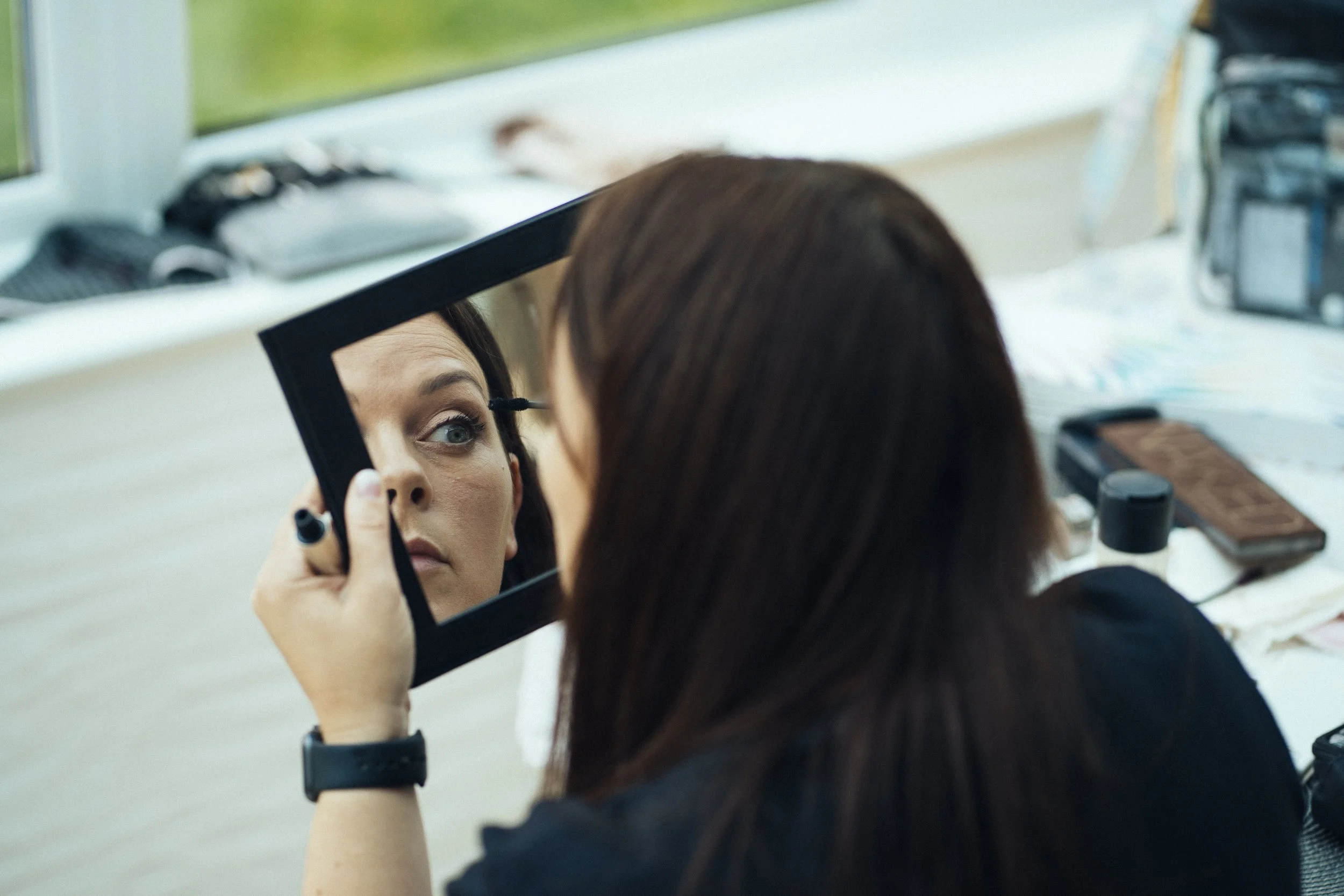 A woman with brunette hair applying eyeliner while looking into a mirror.