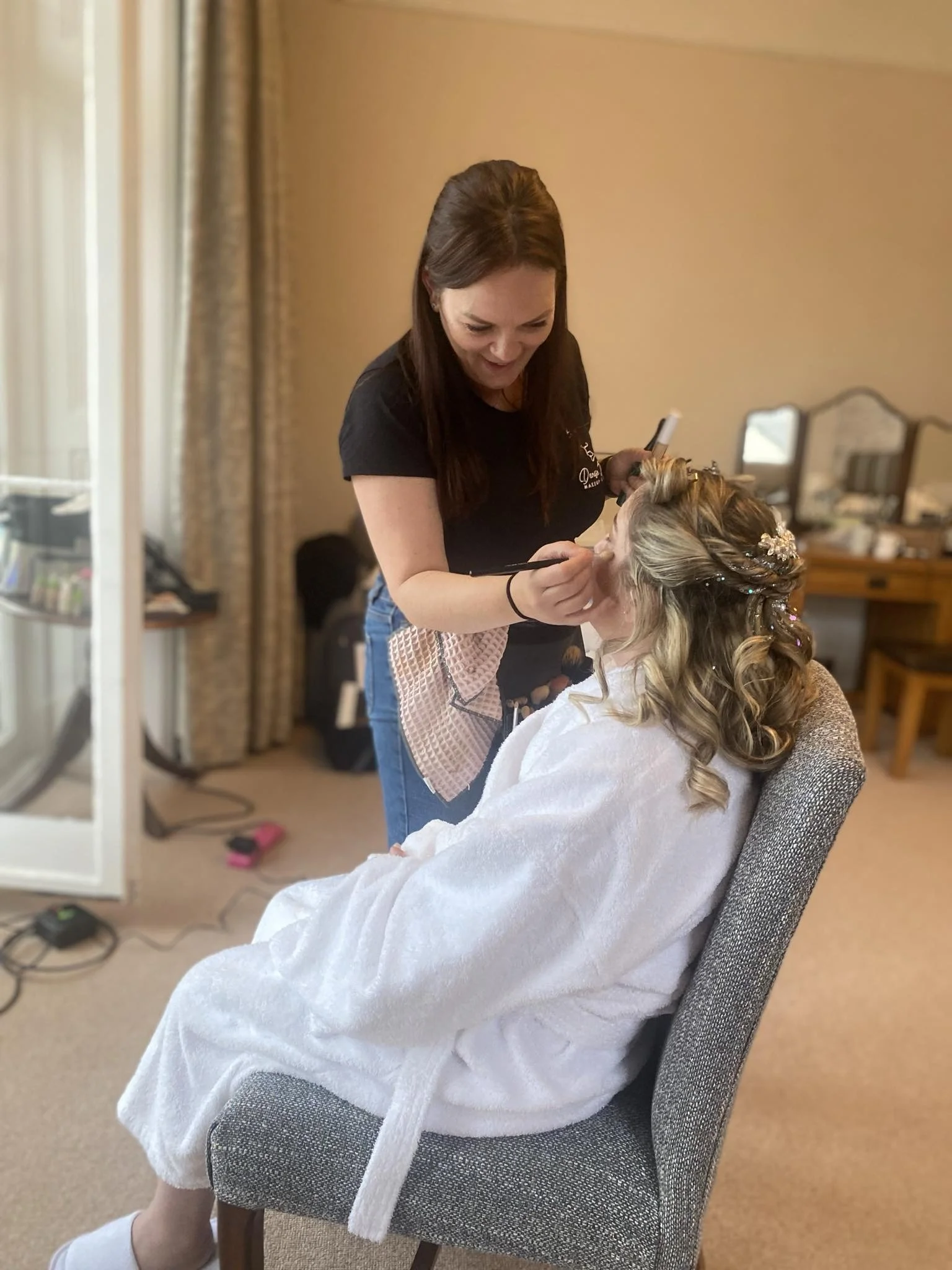 Denise Jackson is applying makeup to a seated bride with blonde, curled hair in a salon or dressing room.