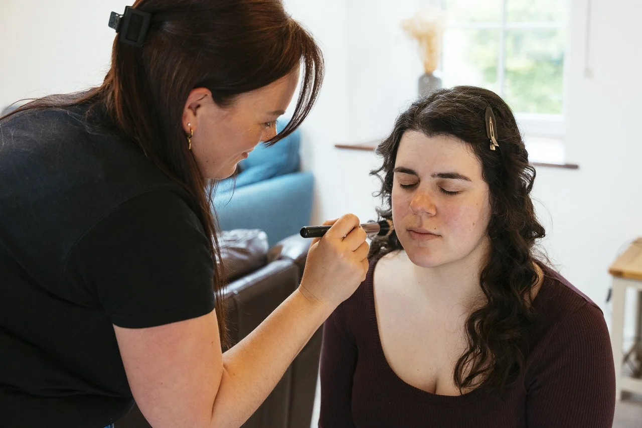 A woman with dark curly hair having makeup applied on her face by makeup artist Denise Jackson in a bright room with a window in the background.