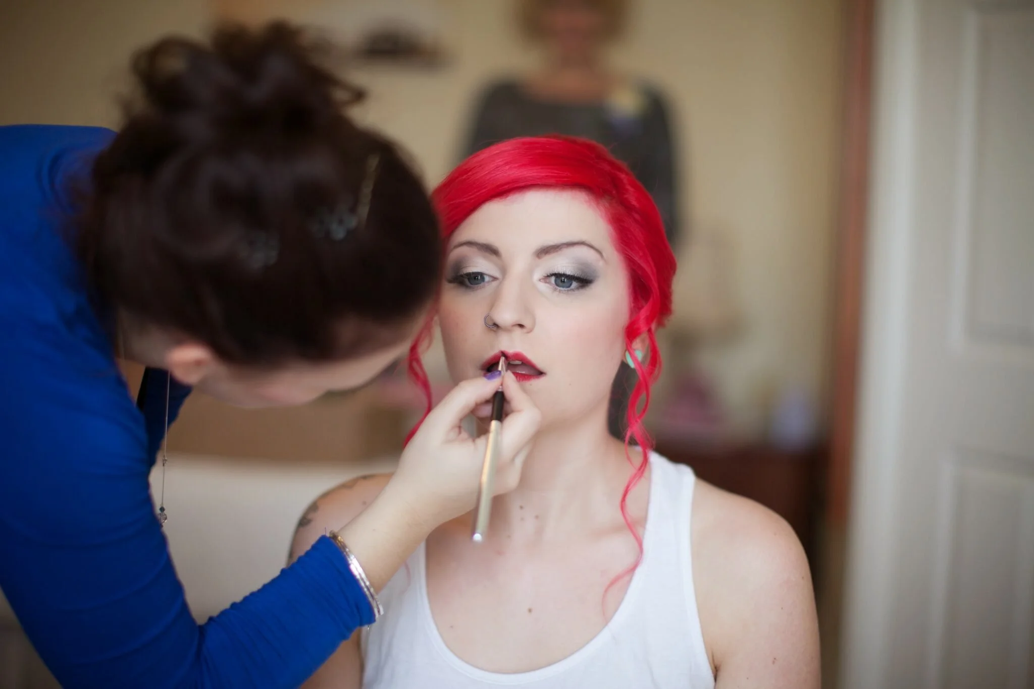 A woman with bright red hair and a white tank top is getting her makeup done by another woman wearing a blue top. The makeup artist is applying lipstick to the woman with red hair.
