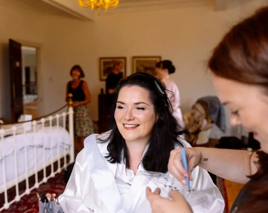 A woman with dark hair smiling as she gets her makeup done in a well-lit room, with three women in the background and a bed on the left side.