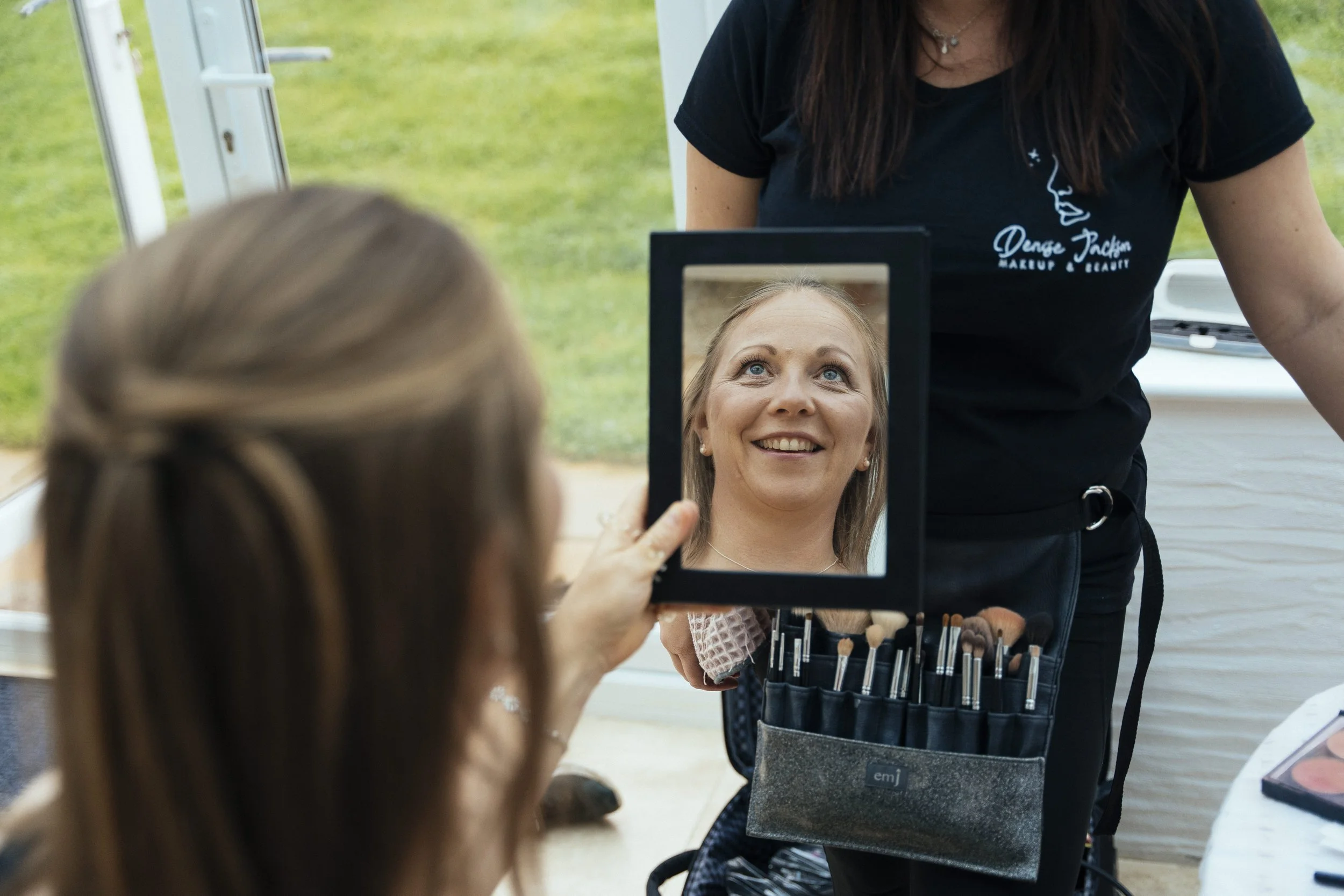 A woman with light eyes and blonde hair looking into a mirror and smiling, with a makeup artist standing behind her. The woman is wearing a white patterned top and earrings. The makeup artist is wearing a black shirt with a logo and has a makeup palette and brushes in front of her. The scene appears to be in a bright indoor or outdoor setting with a green background.