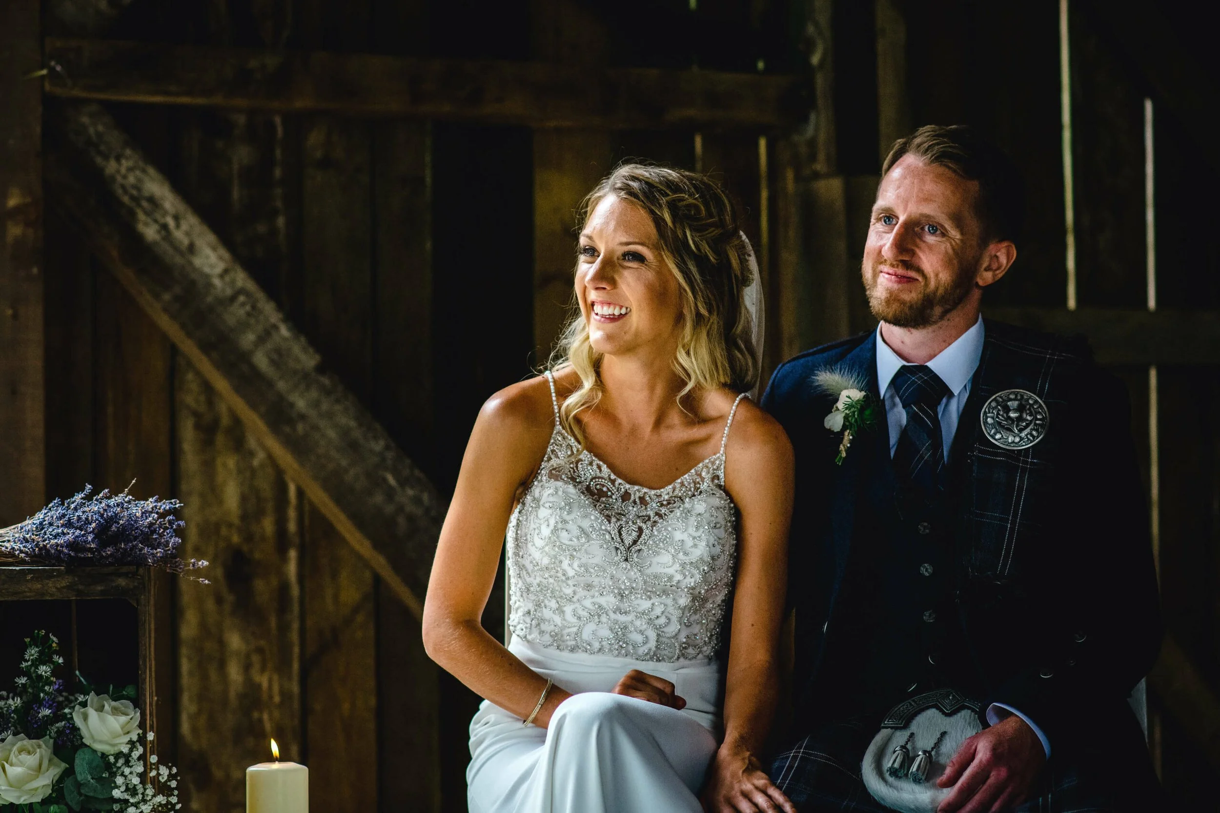 A bride and groom sitting together at their wedding ceremony in a rustic barn. The bride wears a white gown with intricate lace detailing, and the groom wears a traditional Celtic kilt and jacket. There are candles and floral arrangements nearby.