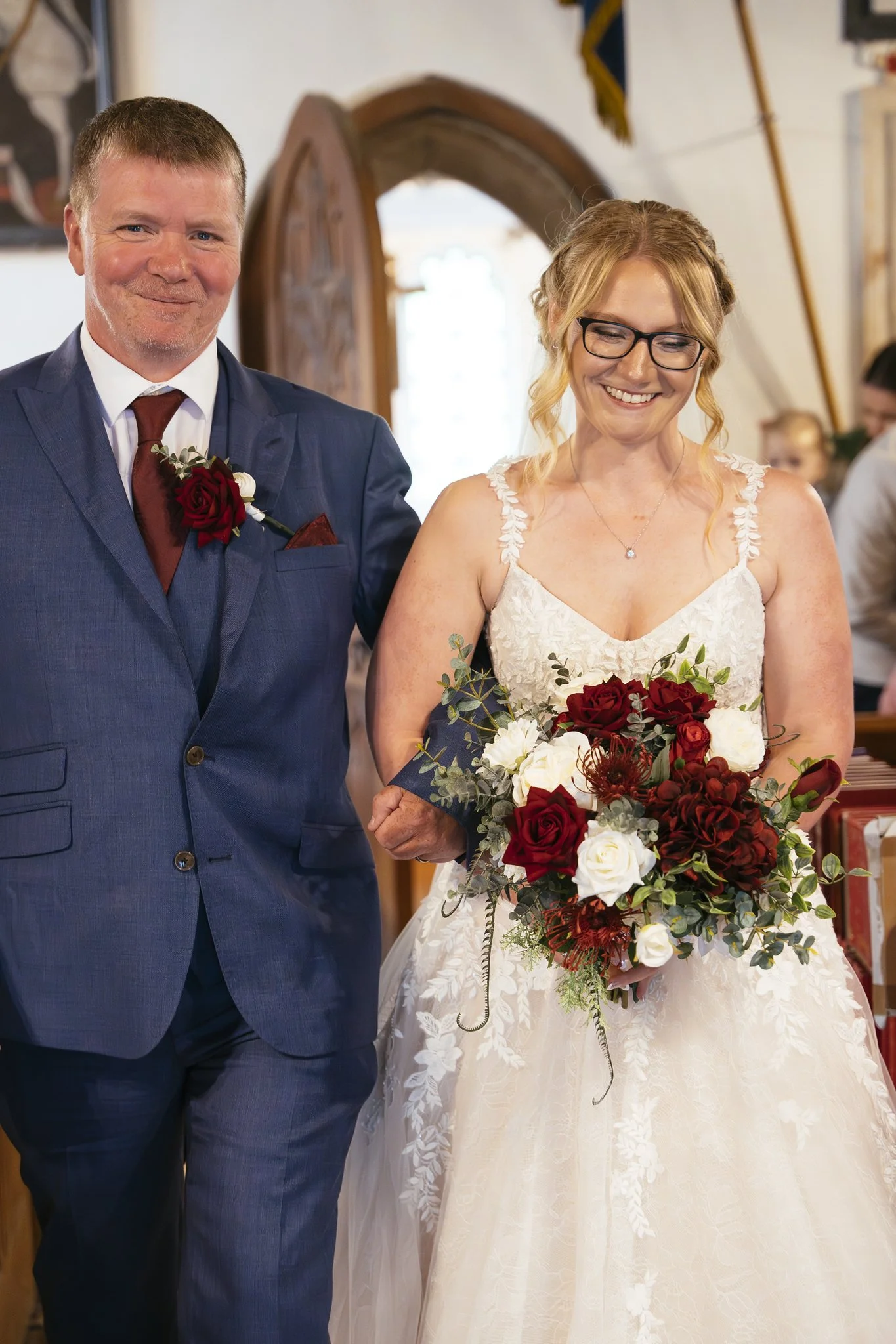 A bride with glasses holding a bouquet of red and white flowers, standing next to a man in a blue suit with a red tie and a red rose boutonniere, in an indoor setting.