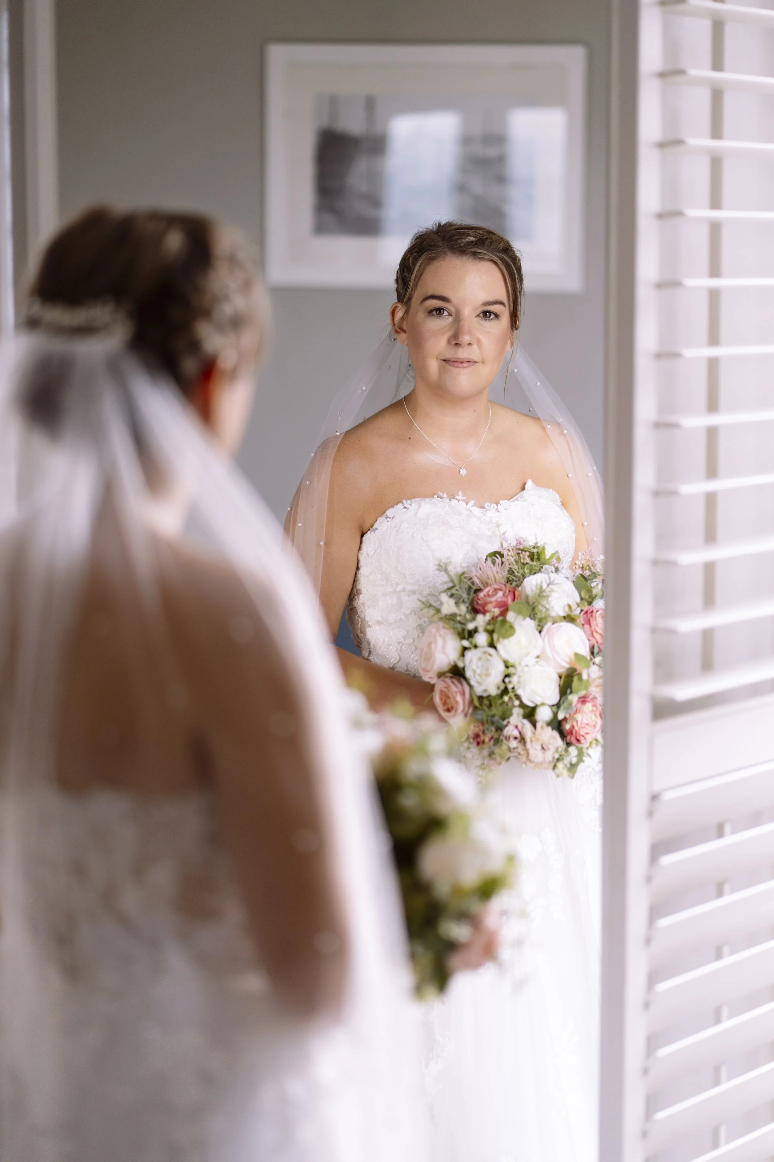 A bride looking at herself in a mirror, wearing a white wedding dress and holding a bouquet of flowers.