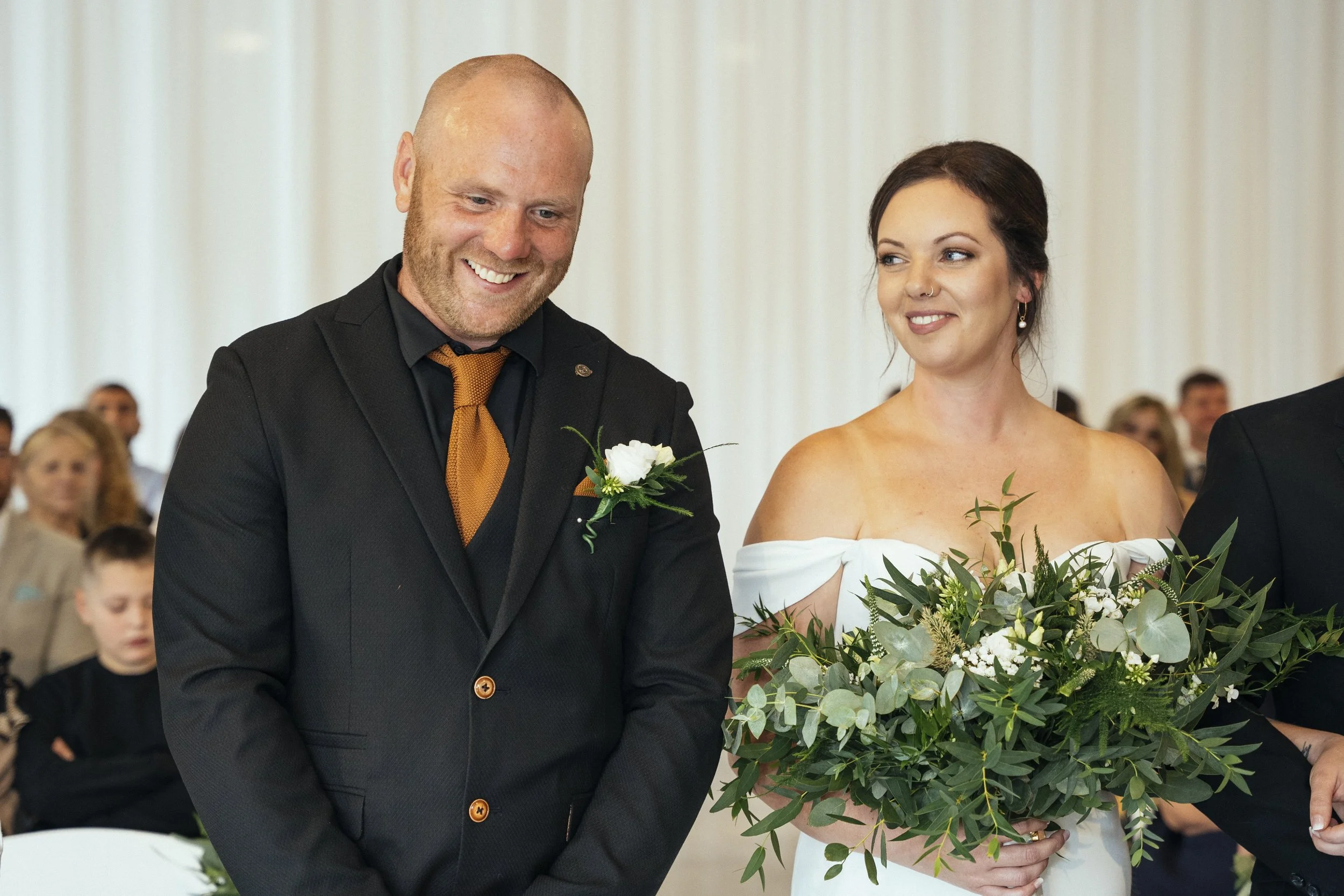 A bride and groom smiling at each other during their wedding ceremony, with the bride holding a large bouquet of greenery and white flowers, and guests visible in the background.