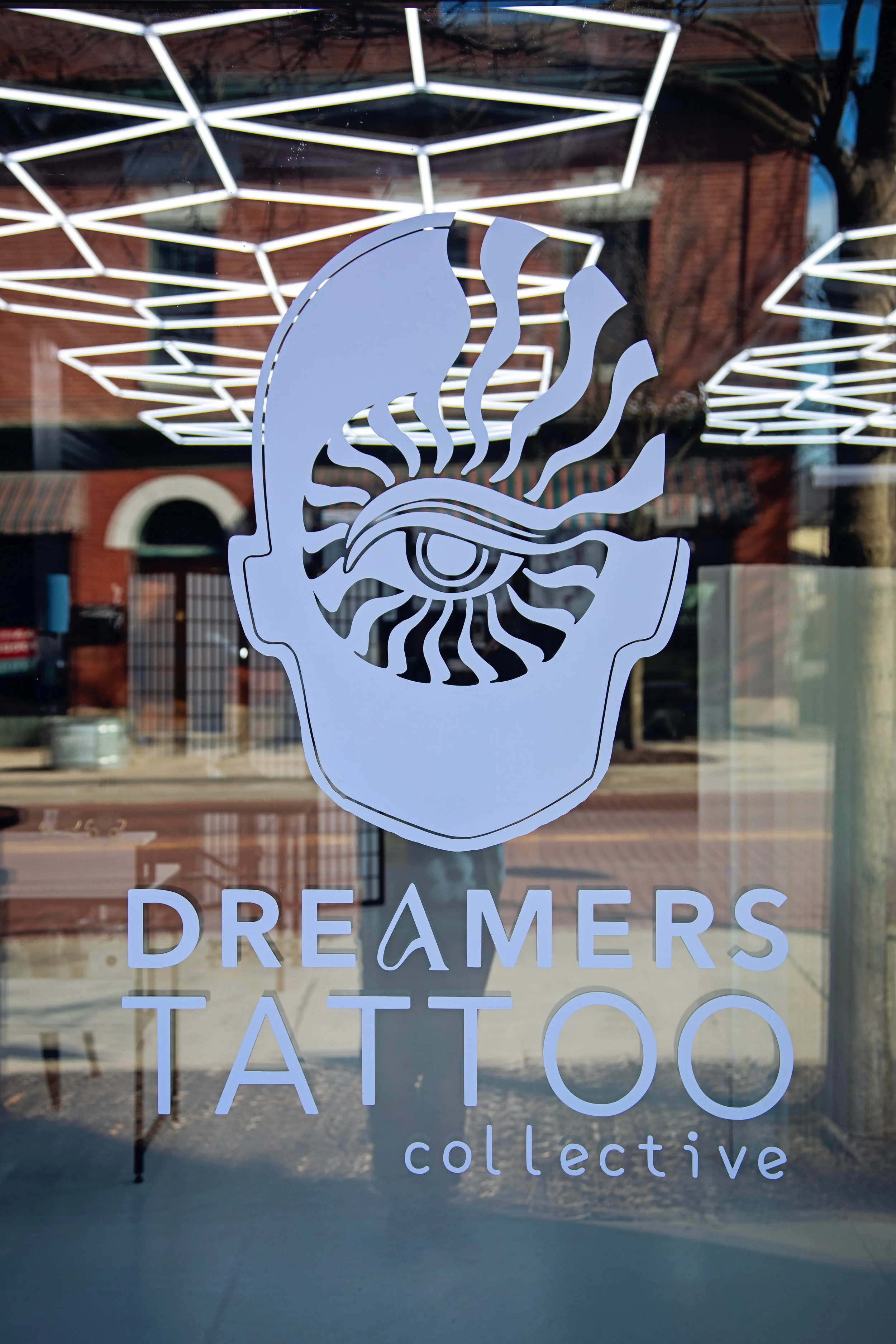 Sign with a stylized face with an eye and abstract facial features above the words "Dreamers Tattoo Collective" on a glass window reflection.