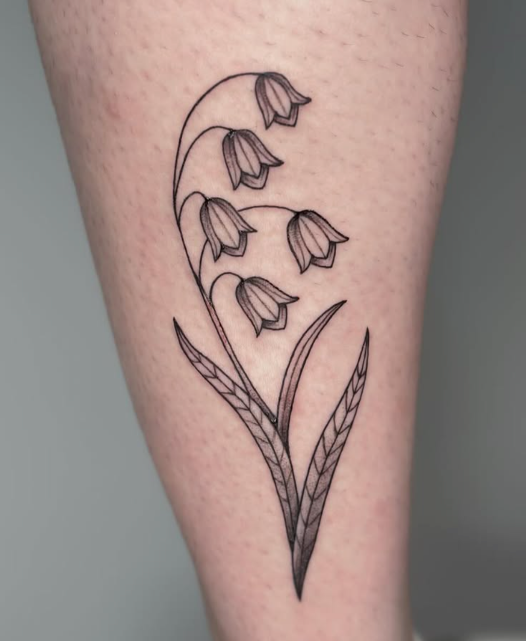 Minimalist black ink tattoo of a lily of the valley flower with multiple leaves on skin.