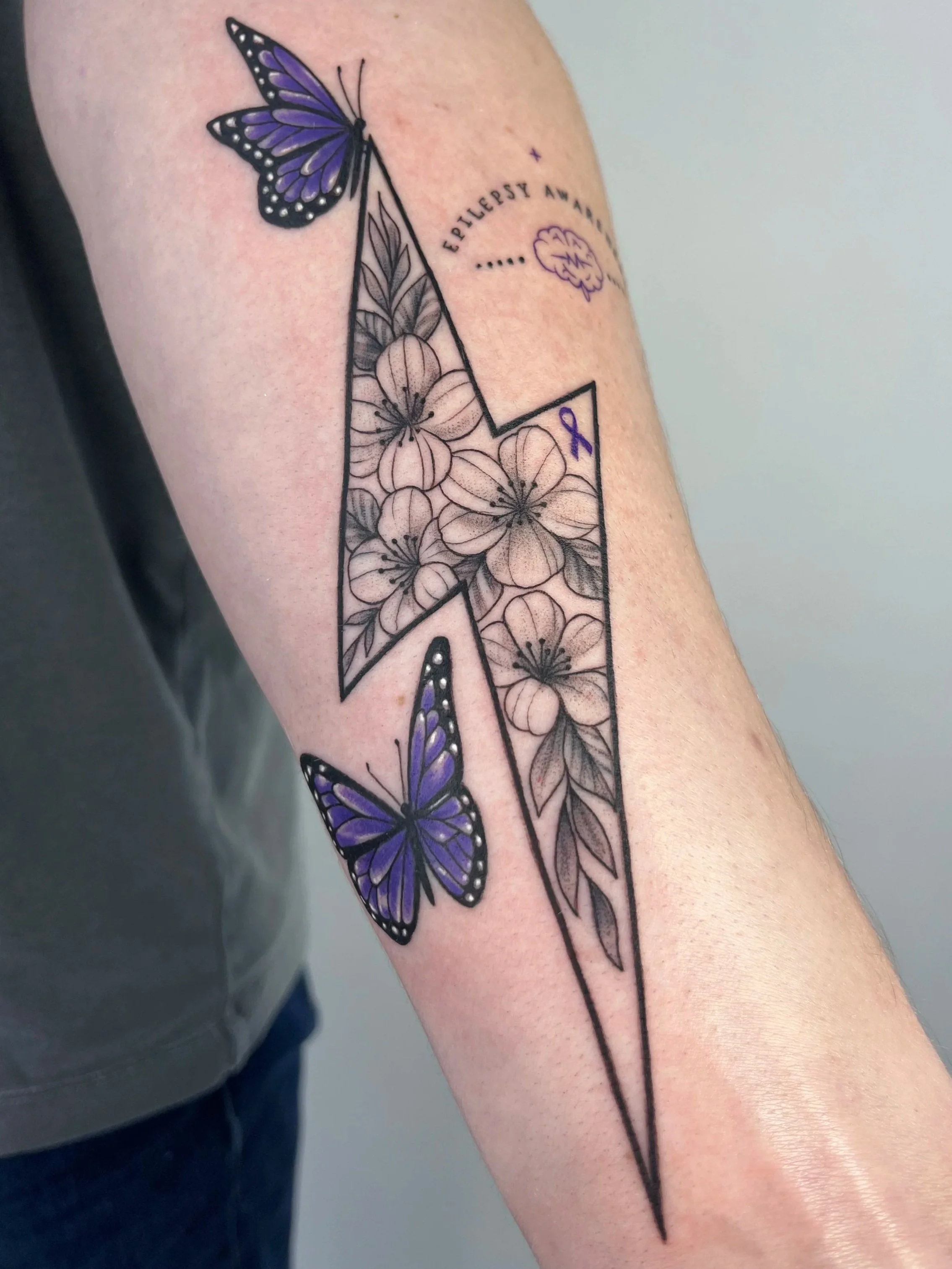 Tattoo of two purple butterflies, one grounded and one flying, with black outlines and shading, along with floral elements, on person's upper arm.