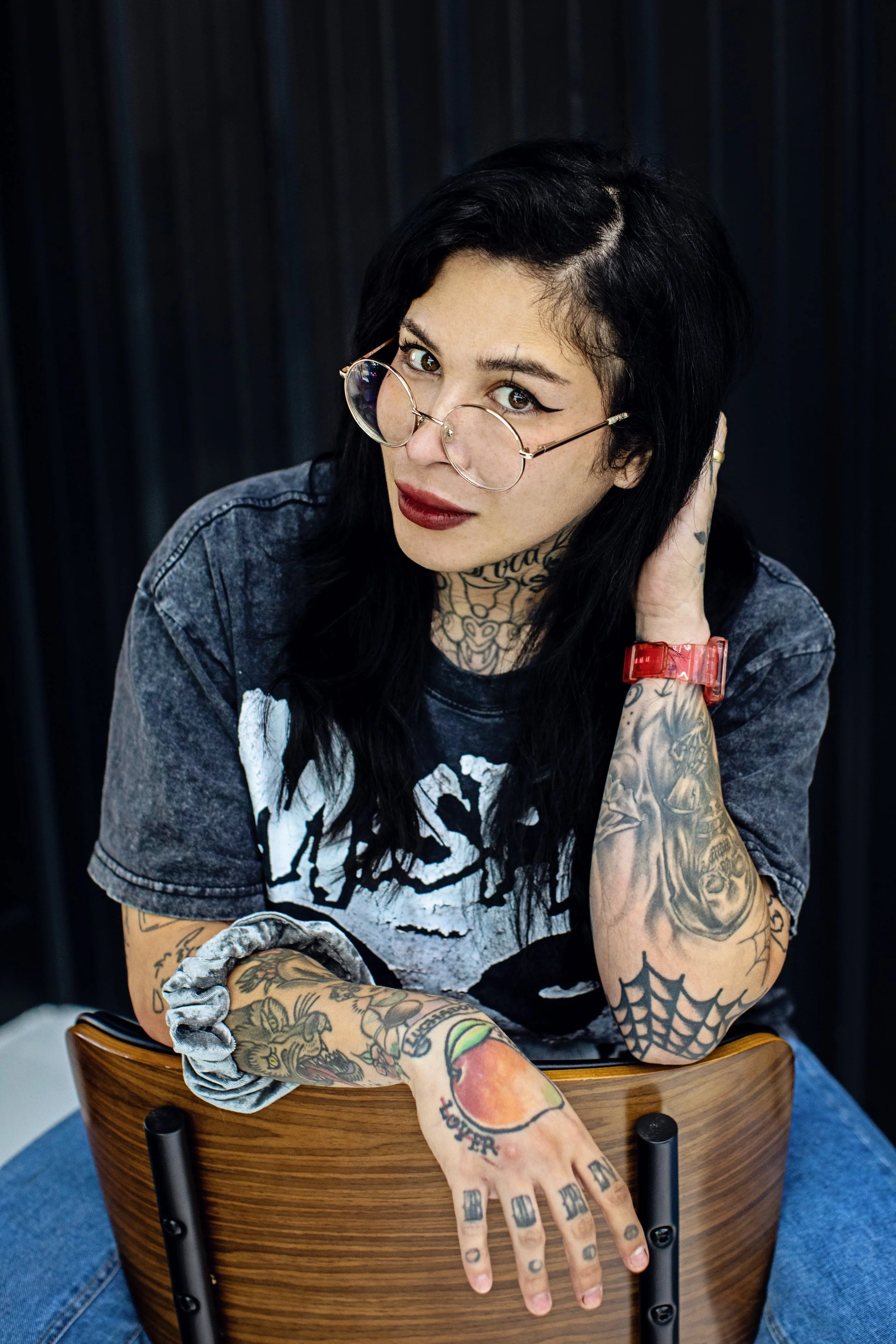 A woman with black hair styled in a vintage bob, wearing glasses with gold frames, makeup including winged eyeliner and red lipstick, and tattoos on her neck and arms. She is smiling and looking down, wearing a black t-shirt with a graphic print.