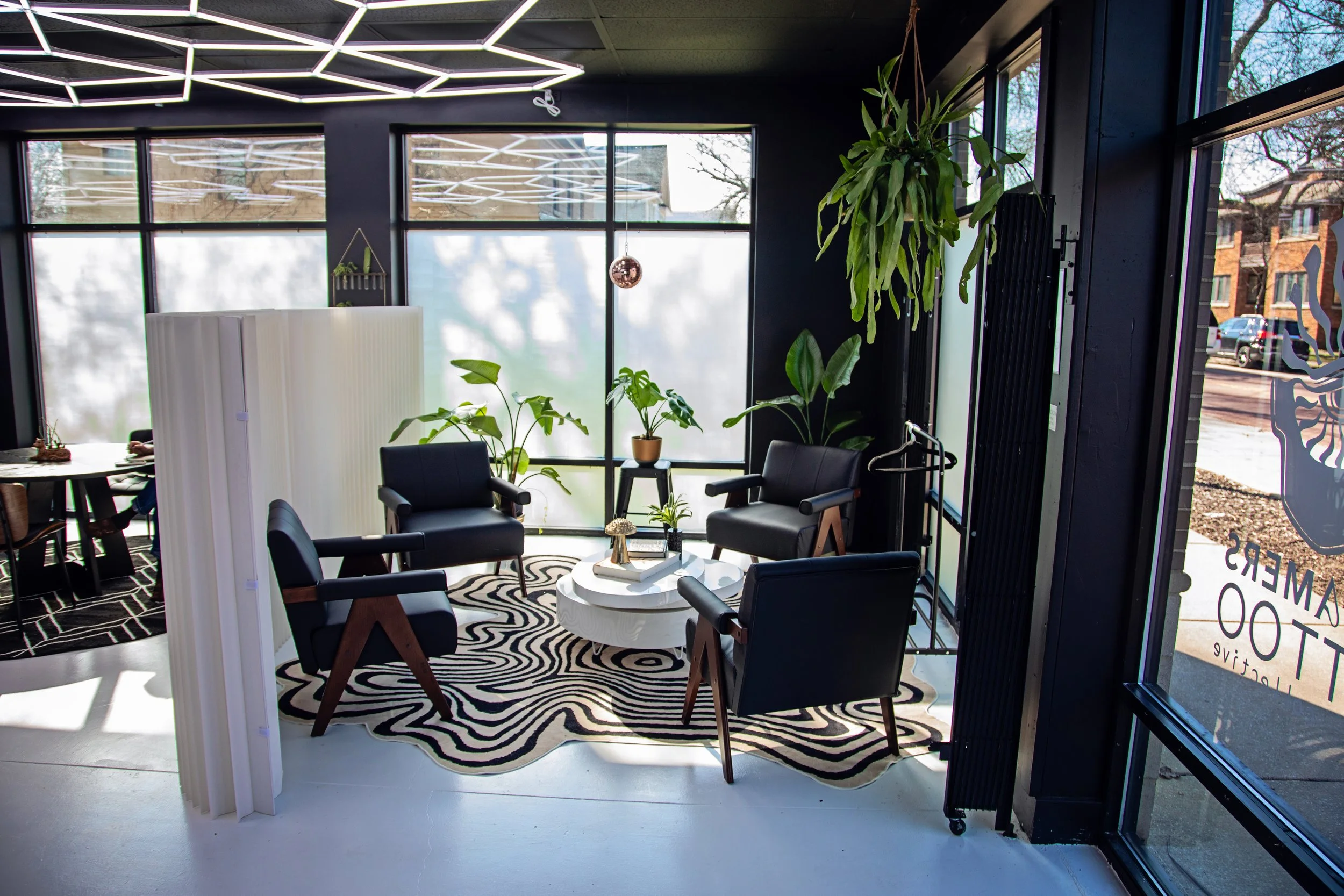A cozy waiting area with four black chairs around a white round table, decorated with plants and a zebra-striped rug, next to large glass windows with black frames allowing natural light.