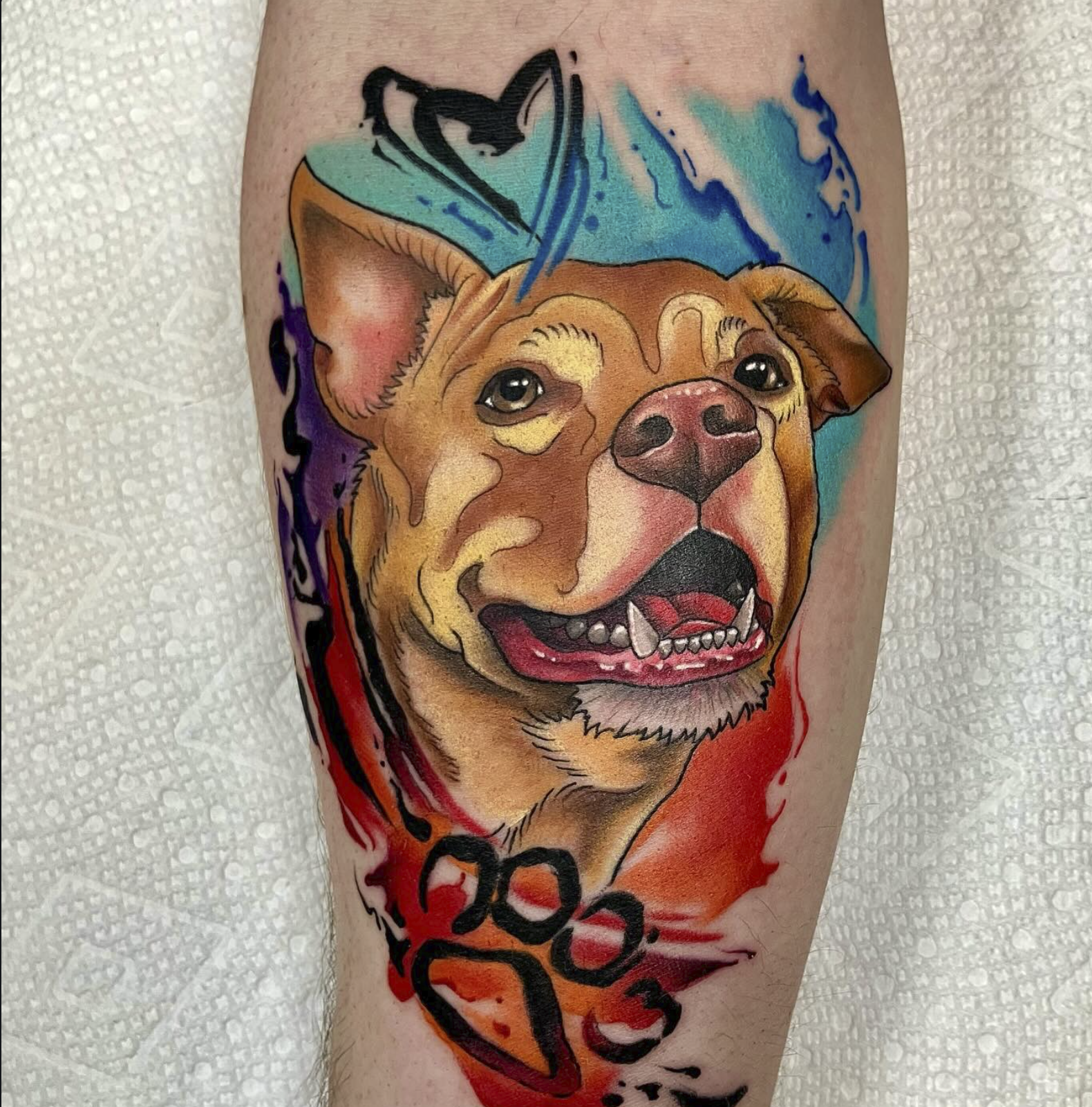 Colorful tattoo of a dog's face with a cheerful expression, surrounded by abstract splashes of blue, red, purple, and black ink with the name 'Loco' written at the bottom.