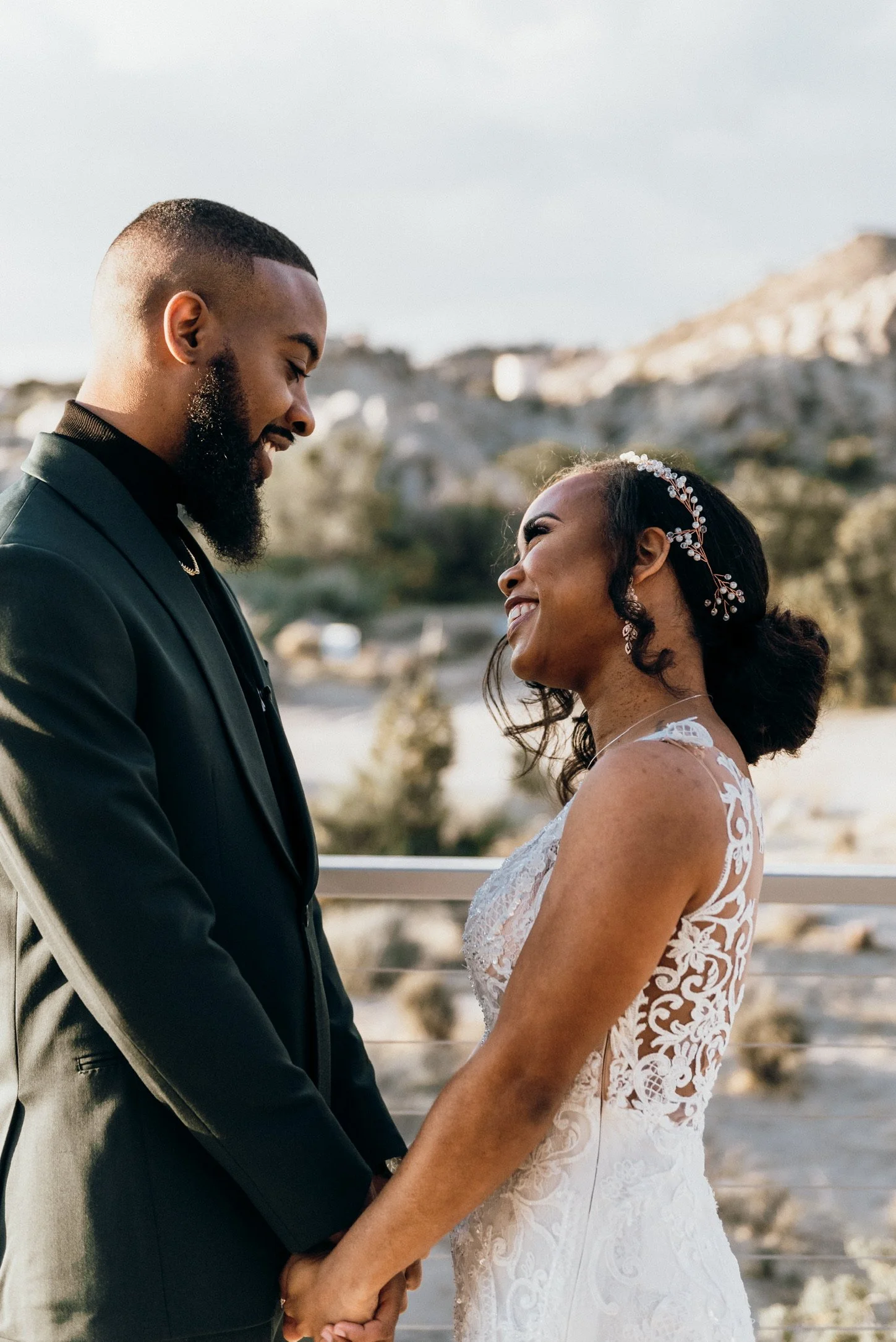 A bride and groom hold hands and smile at each other outdoors during their wedding. The groom wears a black suit, and the bride wears a lace wedding gown with floral embroidery and a hair accessory. Mountains and trees are visible in the background.