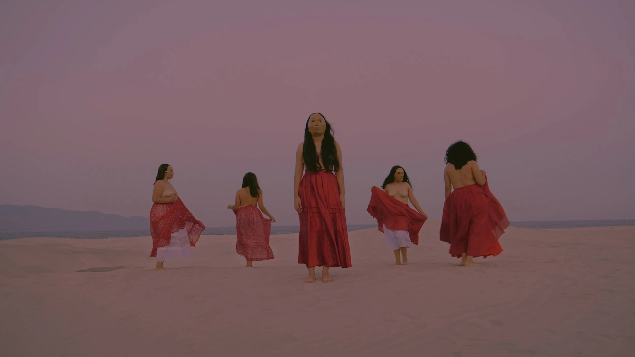 Five women standing barefoot on a sandy desert, wearing long red skirts and topless or with minimal clothing, under a pinkish-purple sky, distant mountains in the background.