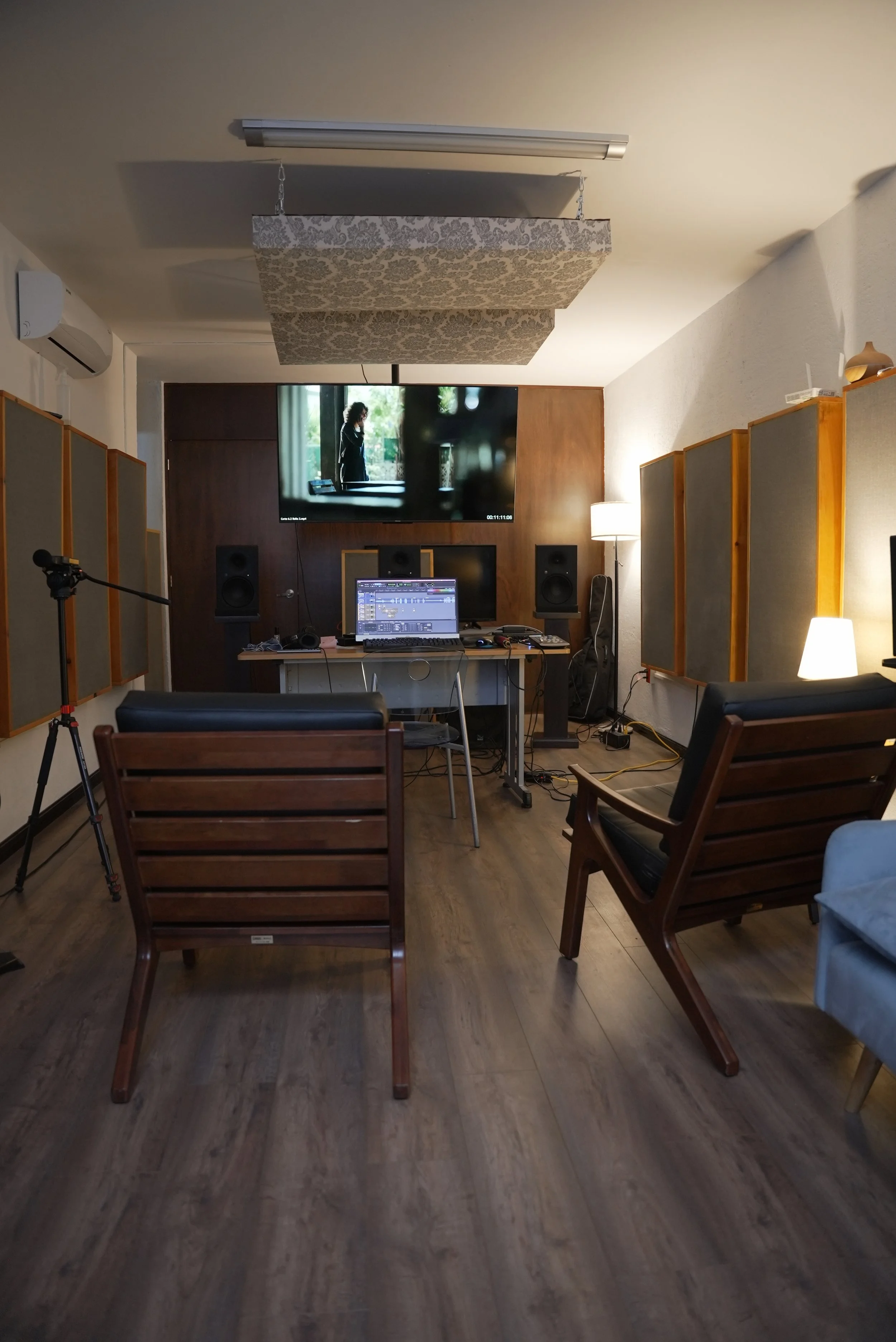Recording studio room with wooden chairs facing a control desk with computer, speakers, and equipment. Wall-mounted acoustic panels, large TV screen, and tripod microphone stand.