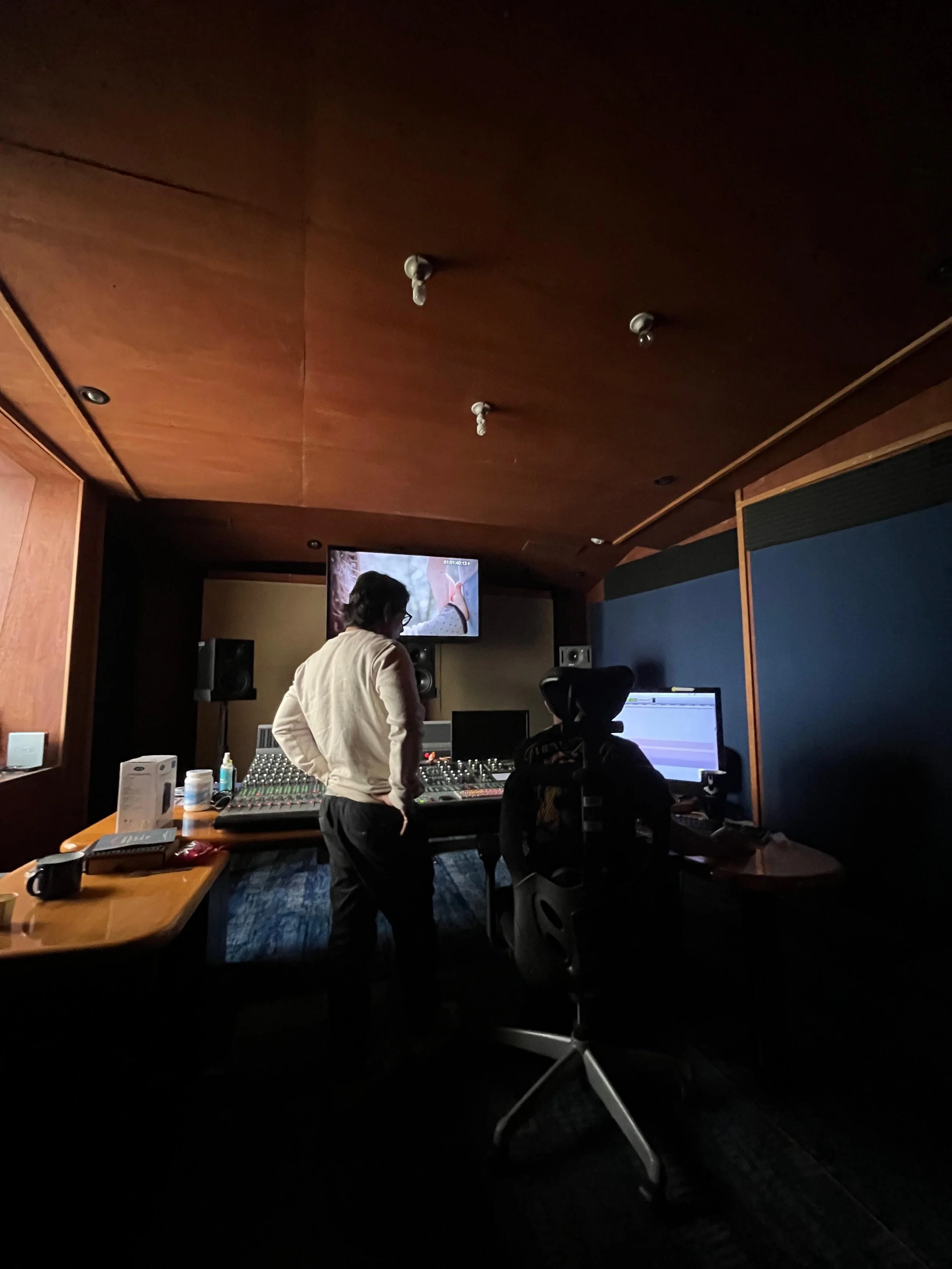 A person standing in a music studio with a mixing console, computer, and recording equipment, facing a large screen on the wall.