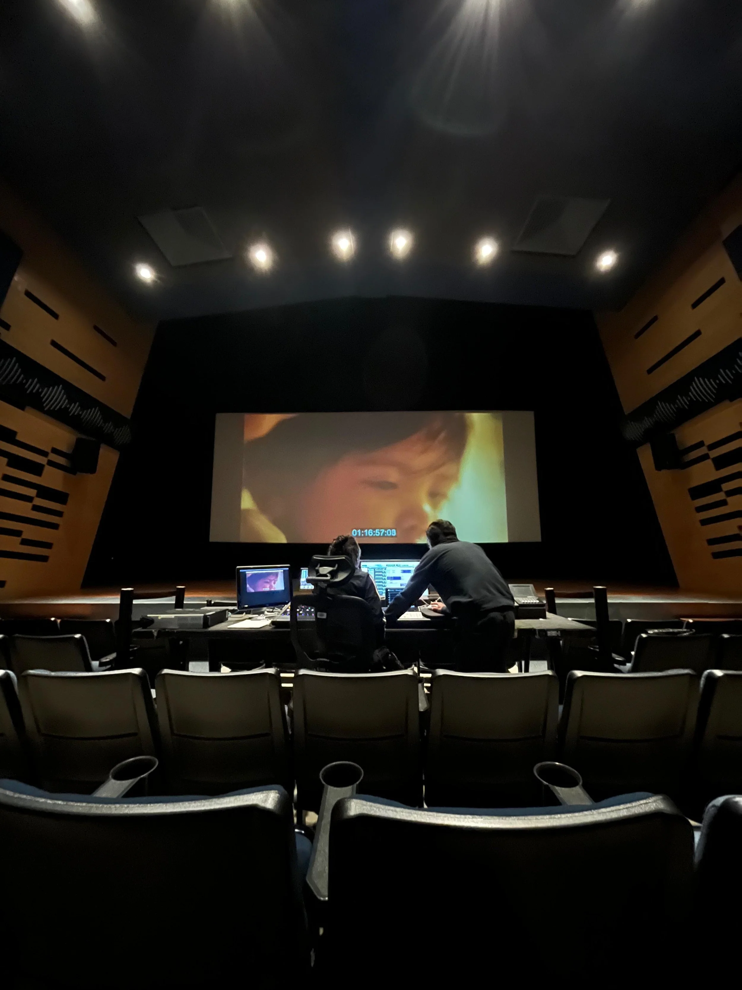 Two people working at a control desk in front of a movie theater screen showing a child's face in a film.
