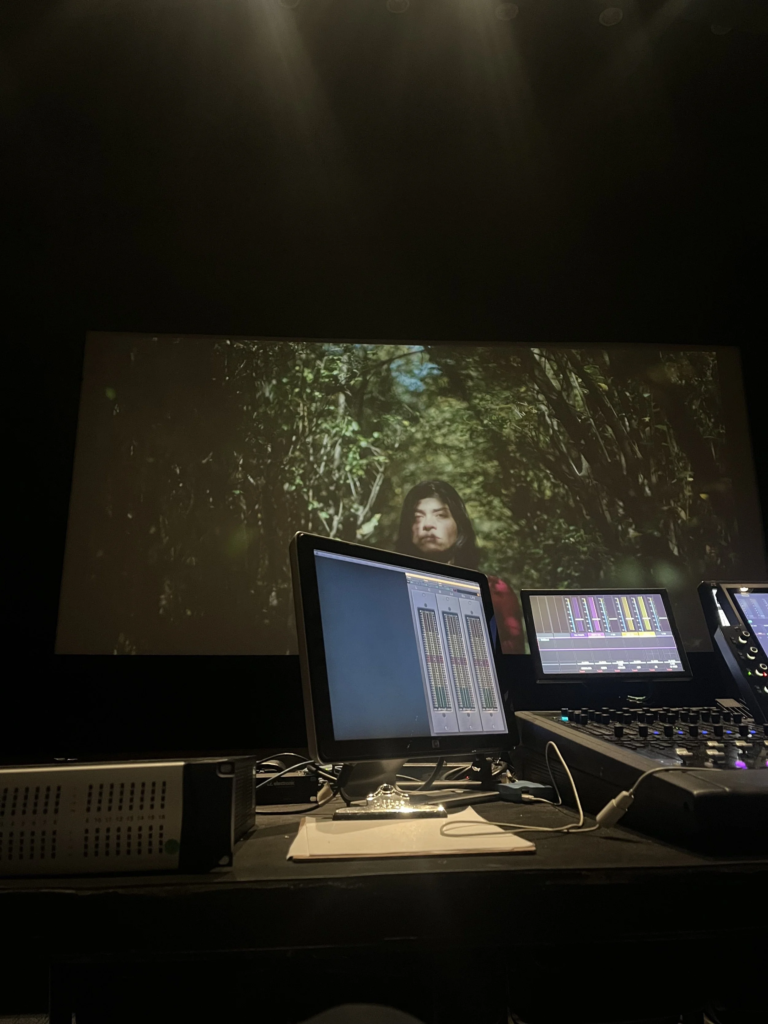 A recording or editing studio with a large screen displaying a woman in a forest, along with multiple computer monitors and audio equipment on a desk.