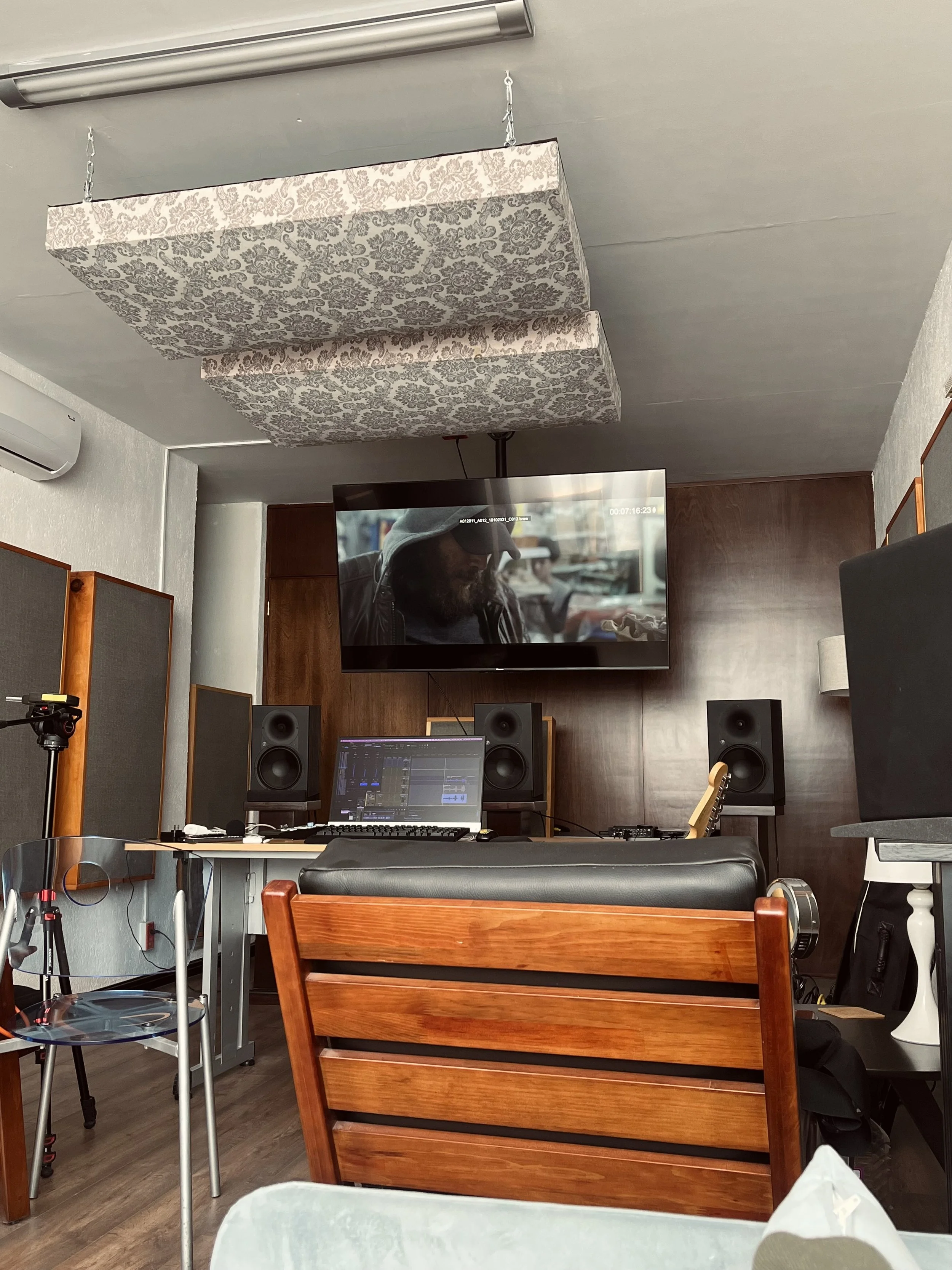 Recording studio with a large TV screen on the wall, showing a man wearing a cap, a computer with audio editing software, two large speakers, acoustic panels, and various equipment.