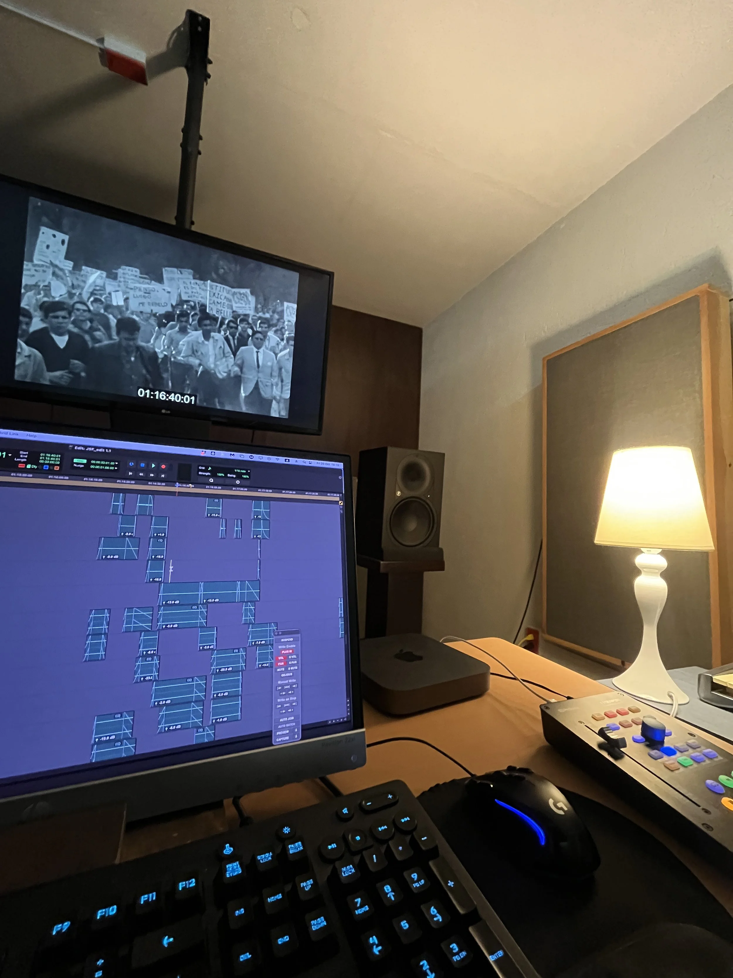 A home recording studio with a computer screen displaying digital audio workstation software, a black keyboard, a gaming mouse, a control surface with colorful buttons, studio monitor speakers, a table lamp, and a video monitor showing a black-and-wh