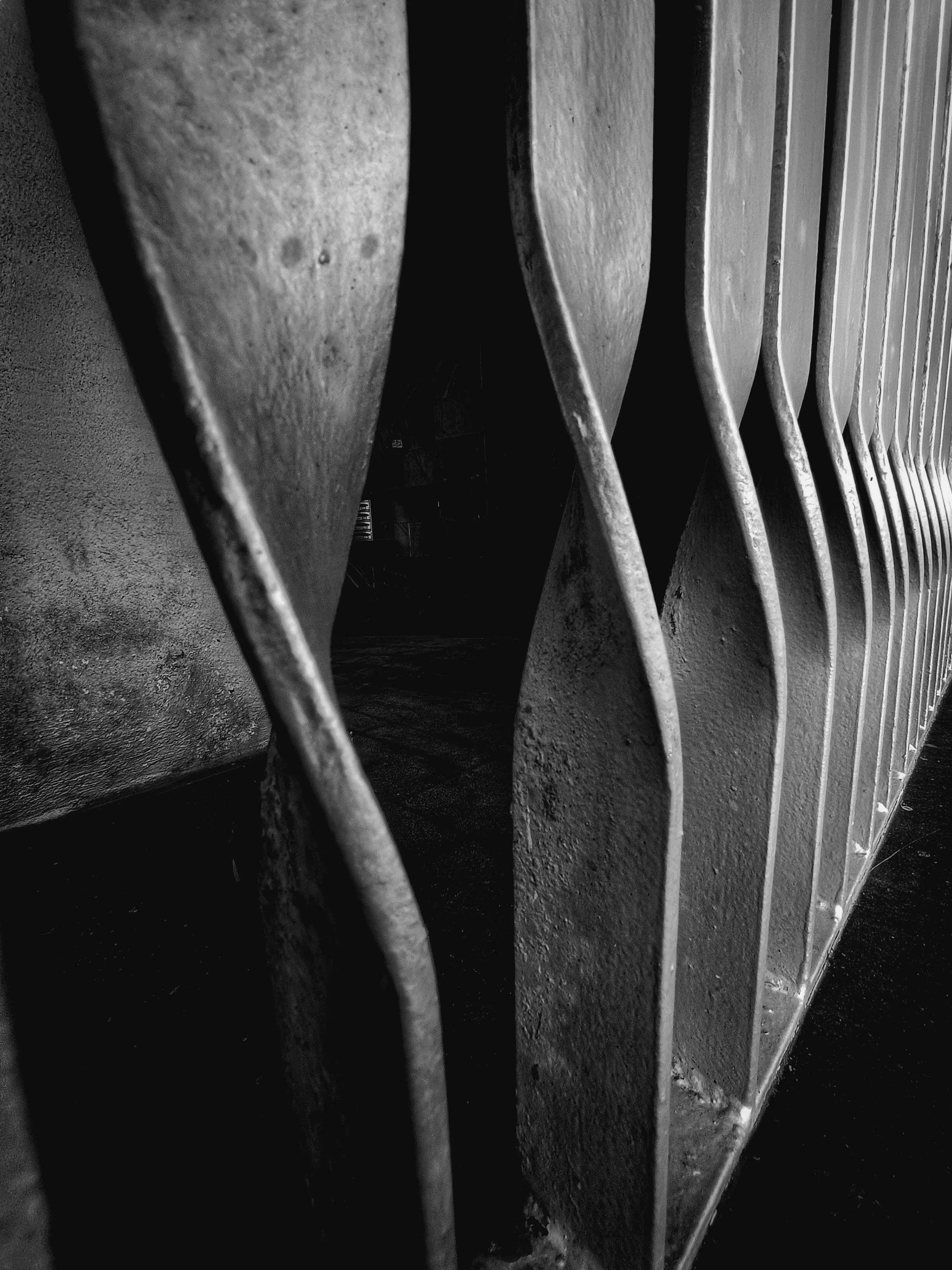 Close-up of vertical metal bars on a railing or fence, with a textured wall behind them and shadows creating contrast, in black and white.