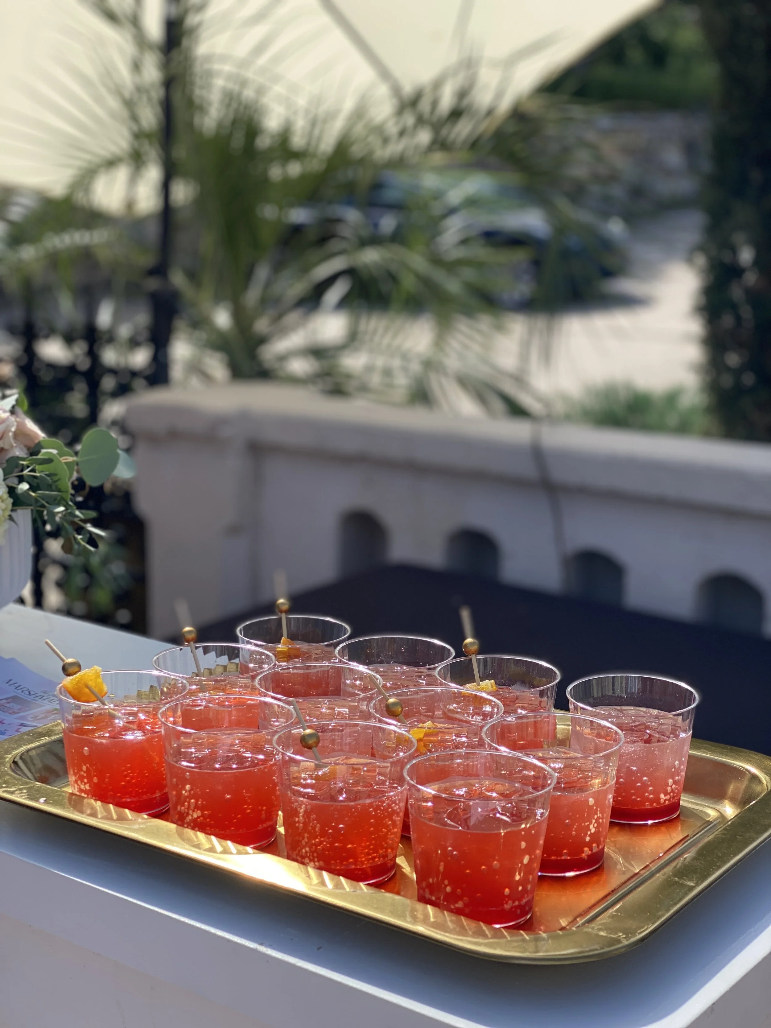 Tray of red-colored drinks with ice and garnished with small skewer snacks, outdoors on a sunny day.