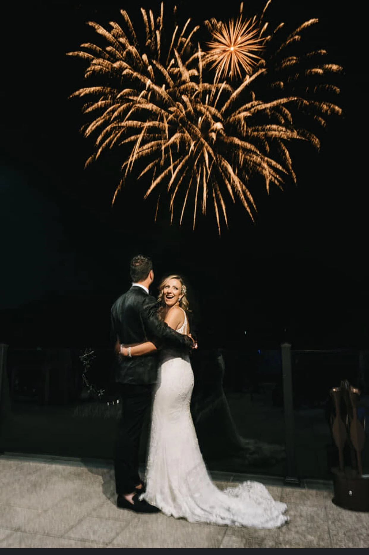 A newly married couple, standing close and gazing at each other, watching fireworks at night. The bride is in a white lace wedding gown, and the groom is in a dark suit.