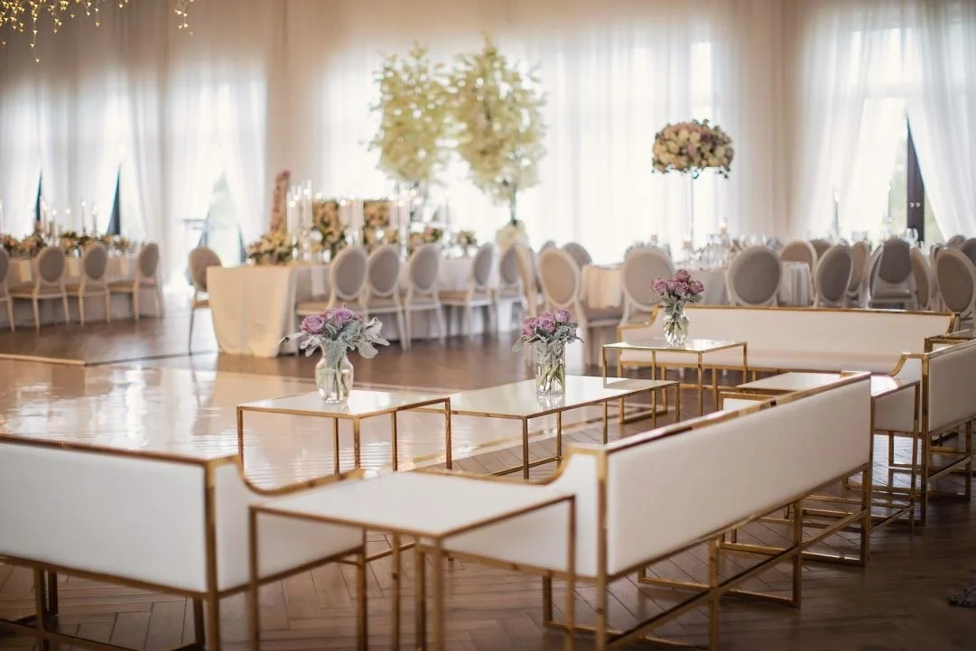 Elegant wedding reception area with white and gold seating, multiple tables decorated with floral arrangements, and a bright, airy ambiance with sheer curtains and large windows.