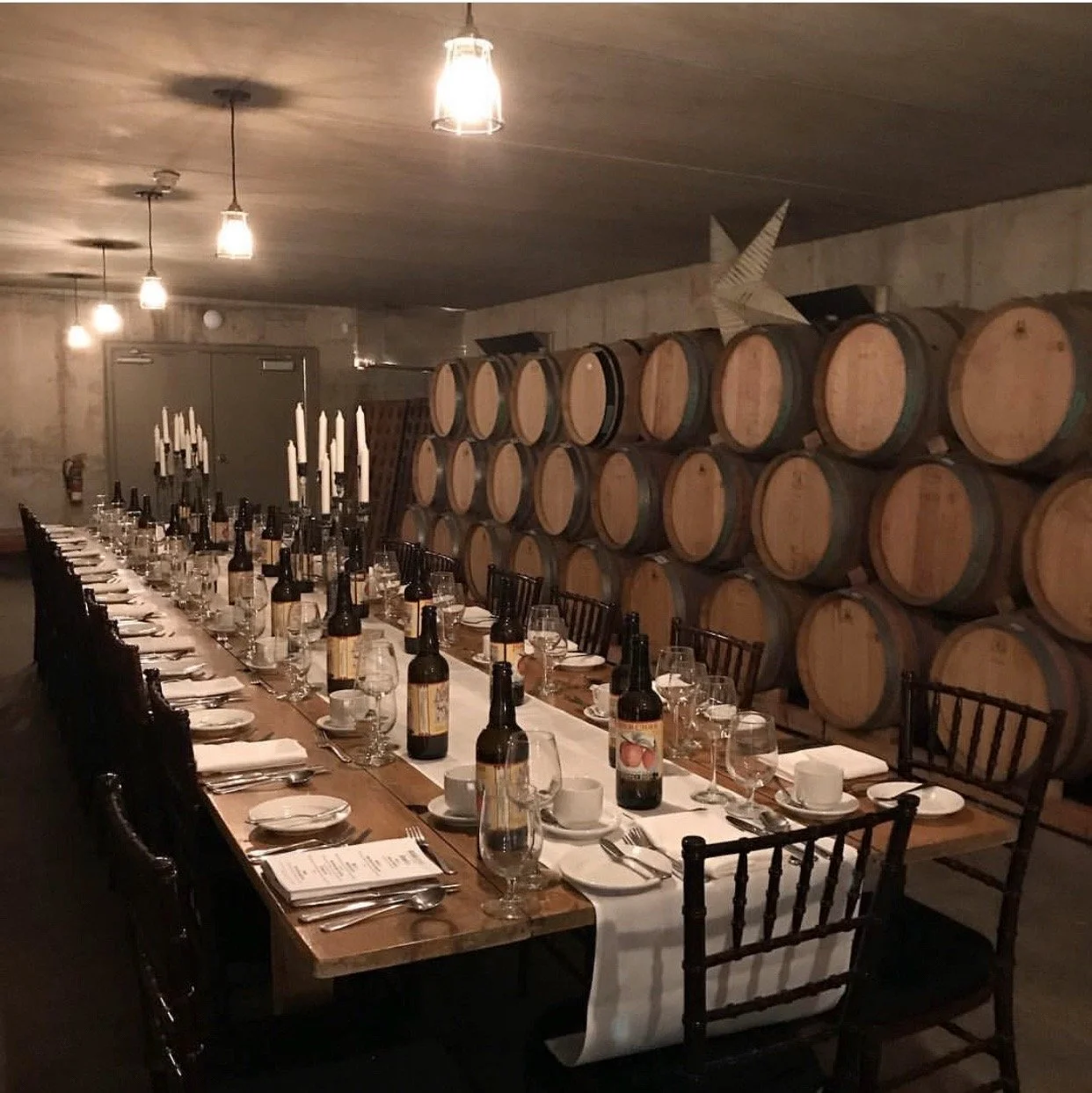 A long dining table set for a dinner, with white tablecloths, plates, glasses, silverware, and bottles of beer. The table is illuminated by several hanging lights. Behind the table are stacked wine barrels against the wall, and a decorative star on t
