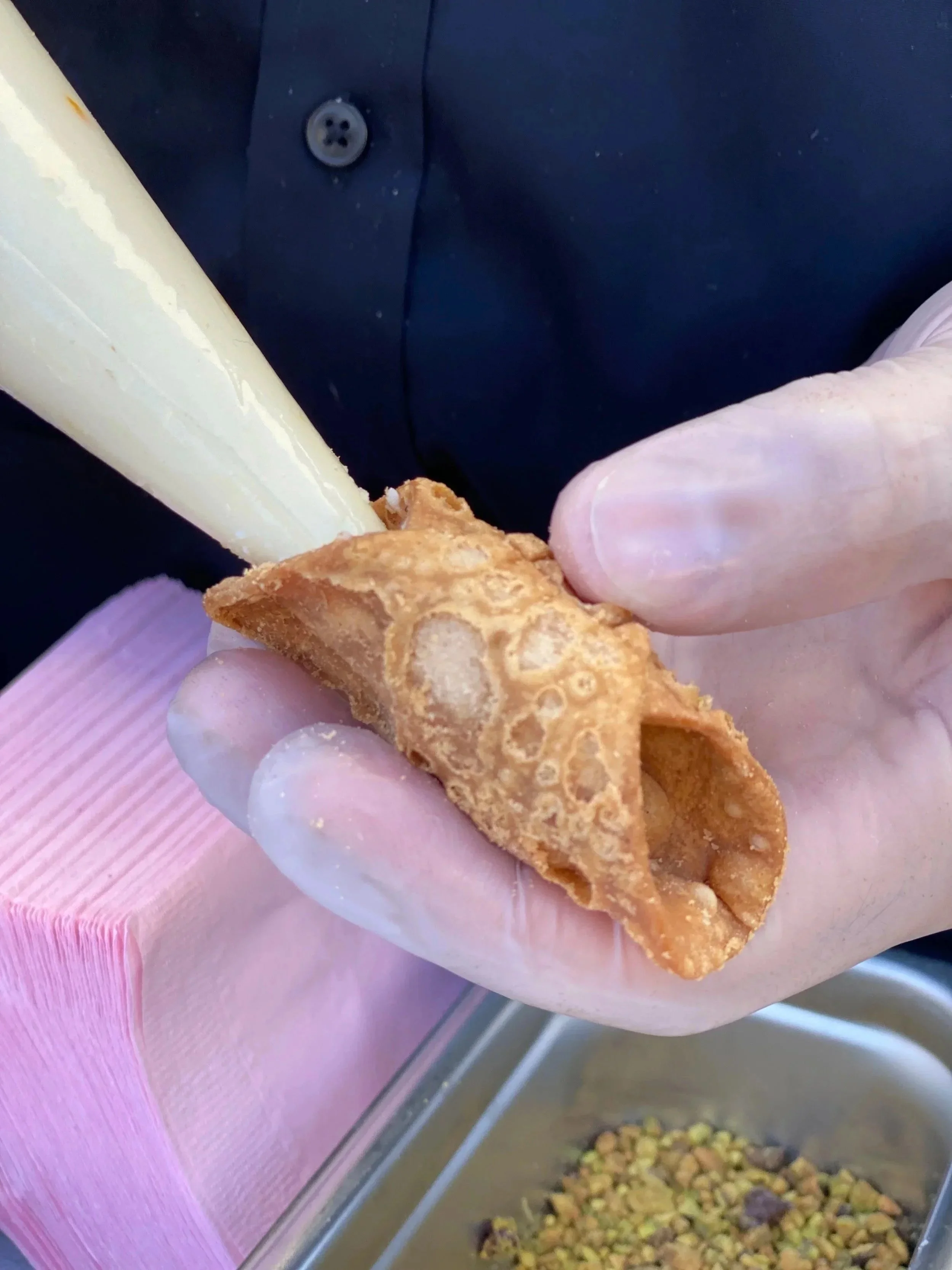 A close-up of a person filling a fried pastry with cream using a piping bag, with a container of chopped nuts in the background.