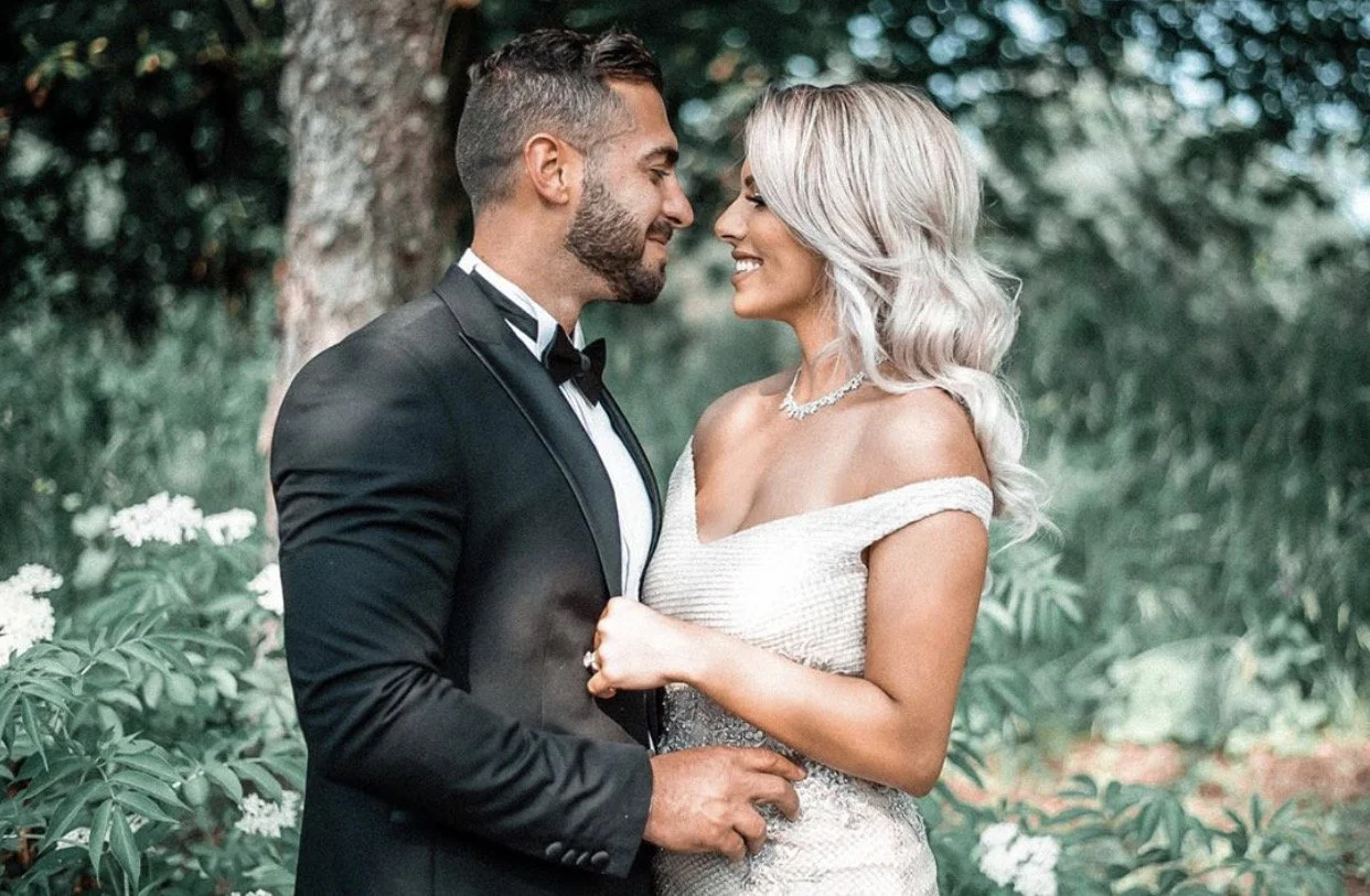 A couple dressed in wedding attire standing outdoors, gazing at each other, with greenery and trees in the background.