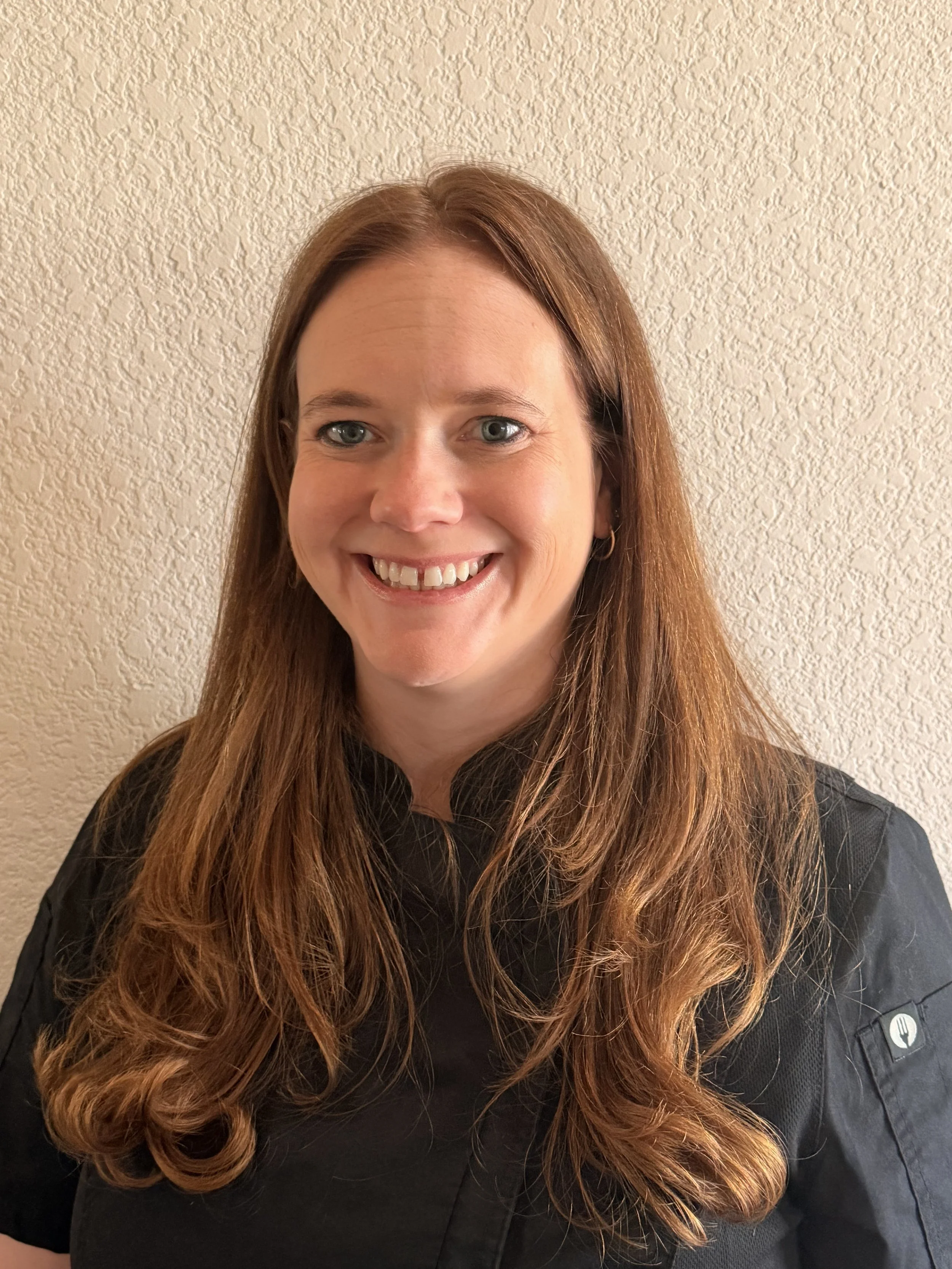 A woman with long red hair and blue eyes smiling, wearing a black chef's coat, standing against a textured beige wall.