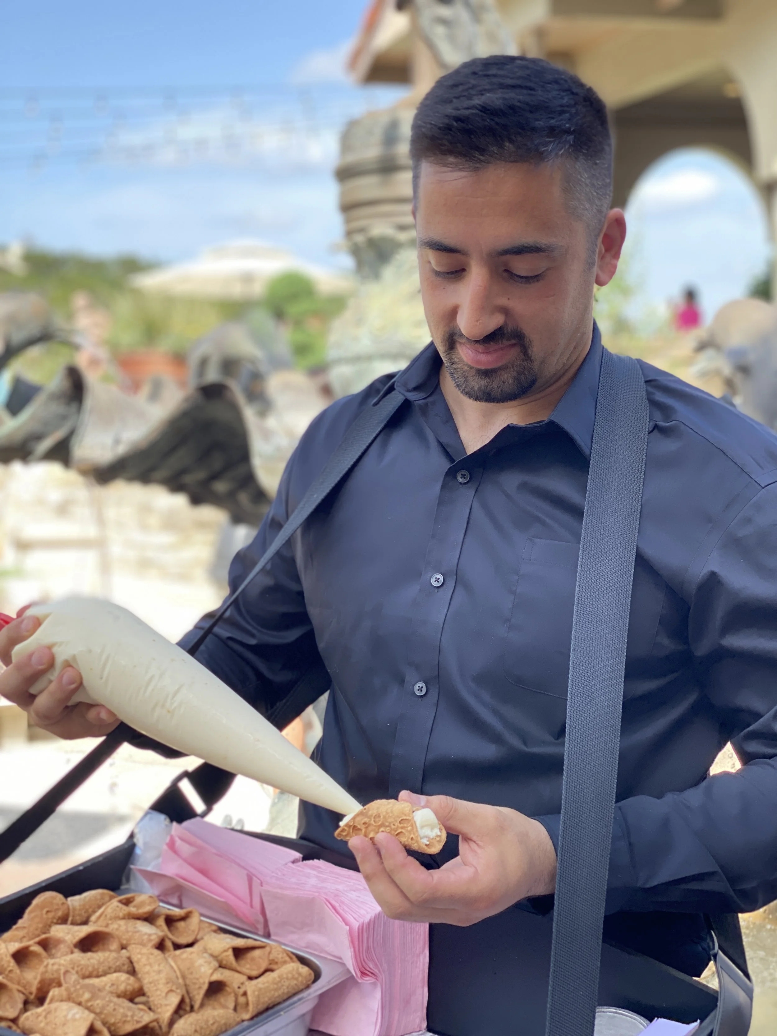 A man in a dark blue button-up shirt holding a cannoli dessert at an outdoor event, with more cannoli pastries on a tray in front of him.