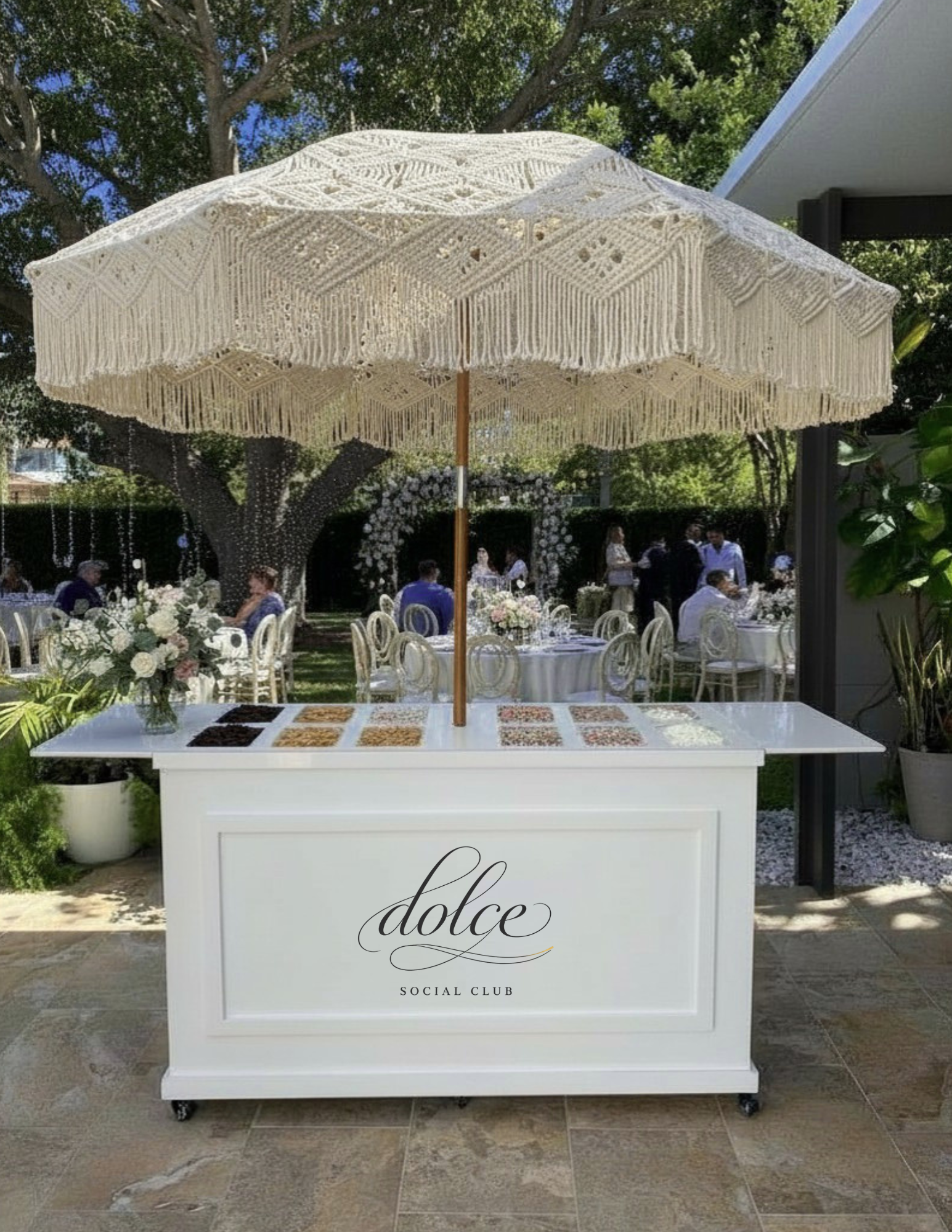 A white cart with a large lace umbrella, decorated with nuts and dried fruit, in an outdoor event setting with tables, chairs, and flower arrangements.
