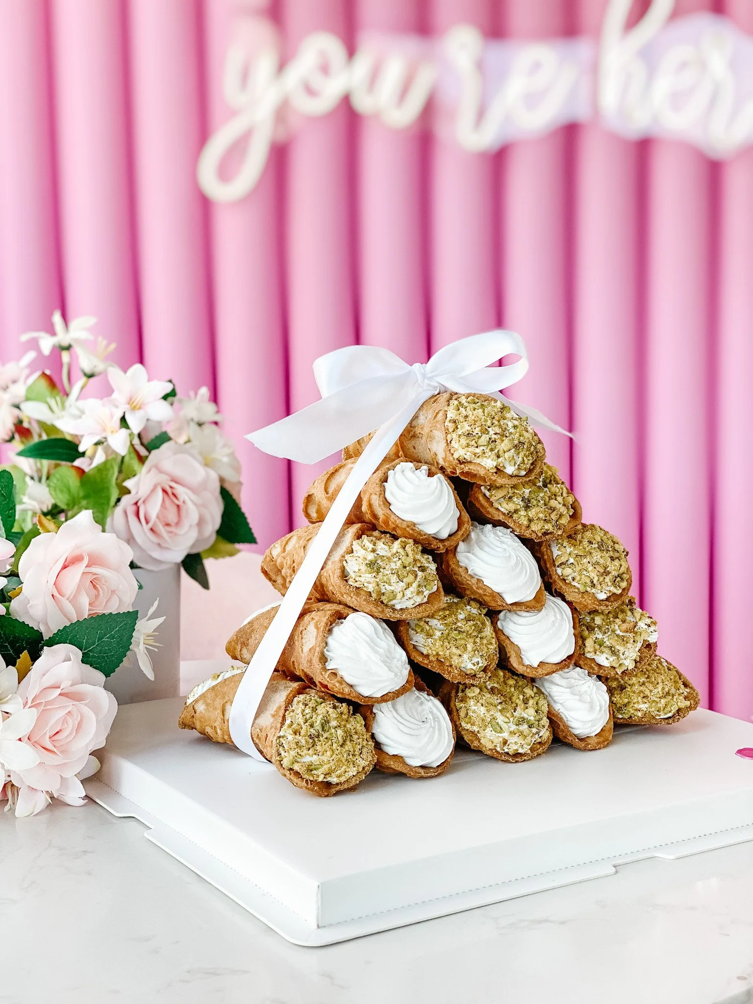 Stack of cream-filled pastry cones with chopped nuts, tied with a white ribbon, on a white board, next to pink roses, with a pink and white striped background and a partial cursive sign.