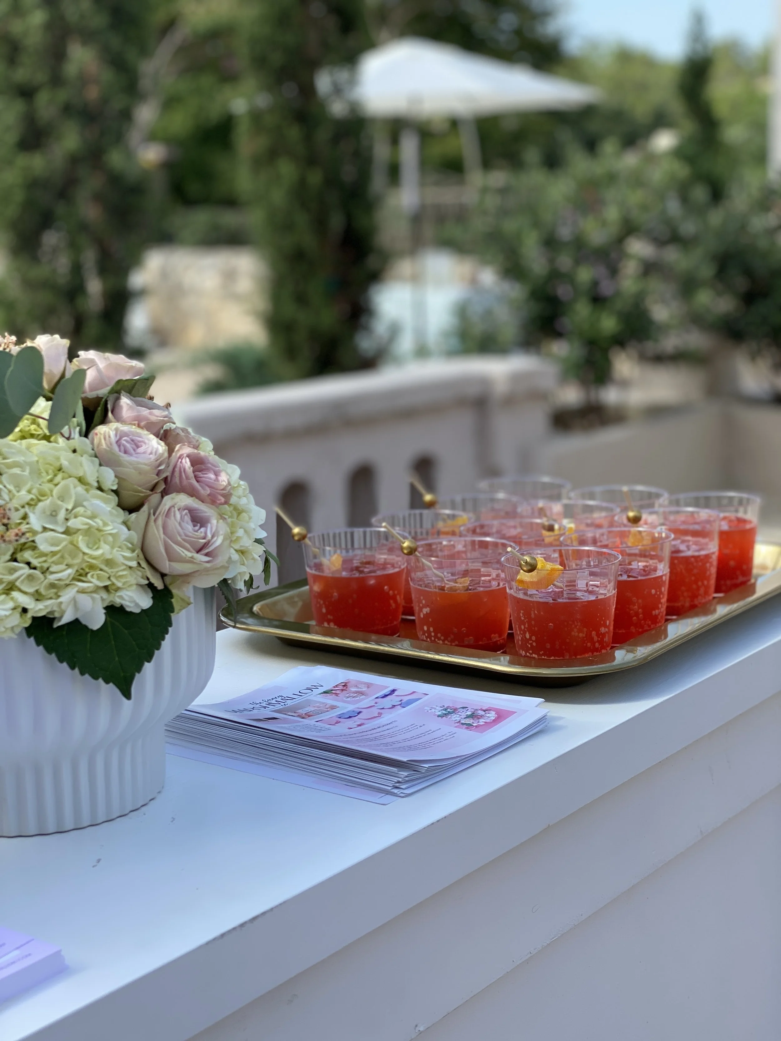 A white table with a bouquet of pink and white roses and hydrangeas in a white vase, and a tray of red drinks with yellow fruit garnishes and small skewers. There are also some pamphlets or brochures on the table.