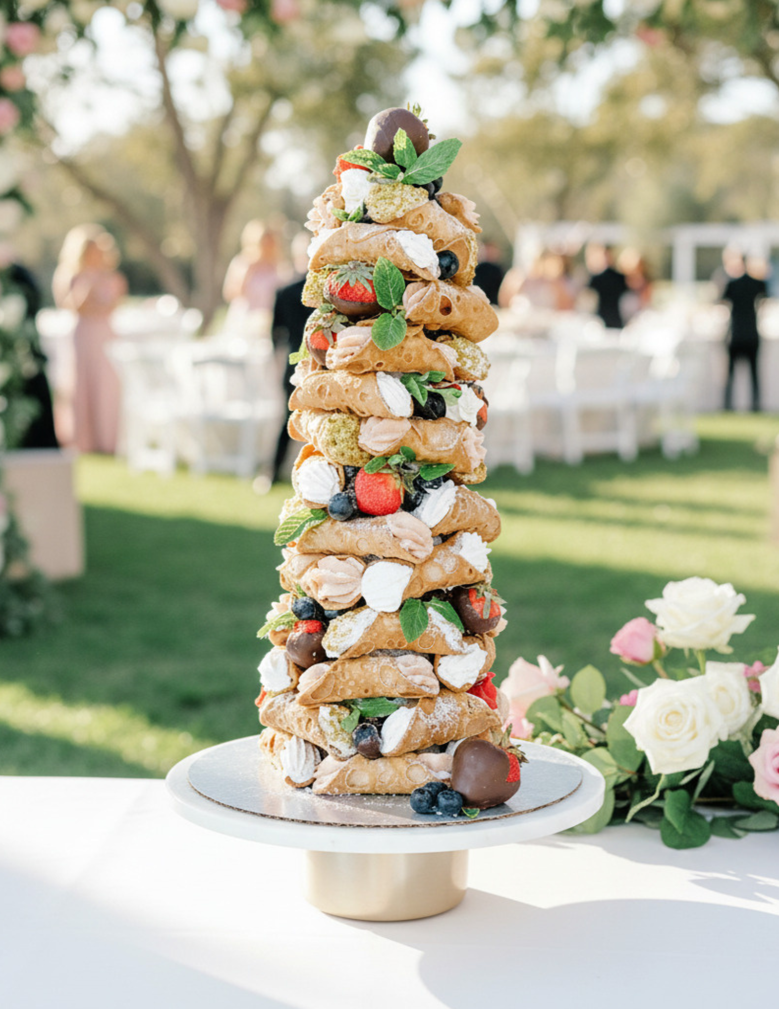A tall, layered dessert resembling a pastry tower, decorated with various fruits and herbs, set outdoors with blurred people and greenery in the background.