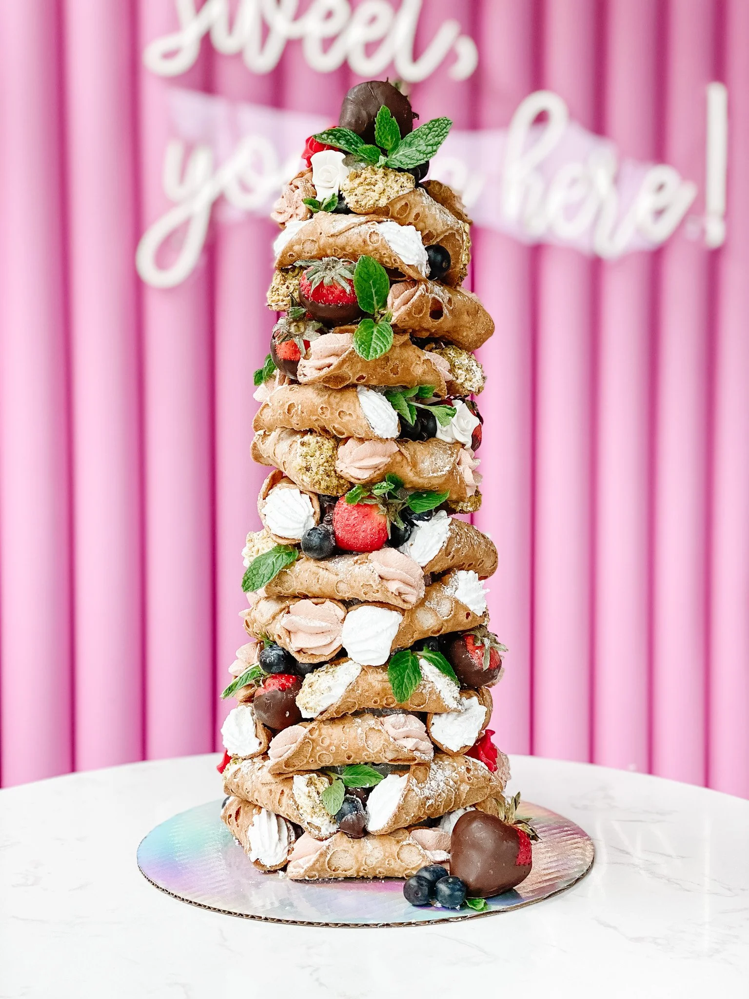 A tall dessert tower made of ice cream cones filled with various berries, chocolate, and whipped cream, decorated with mint leaves against a pink background.