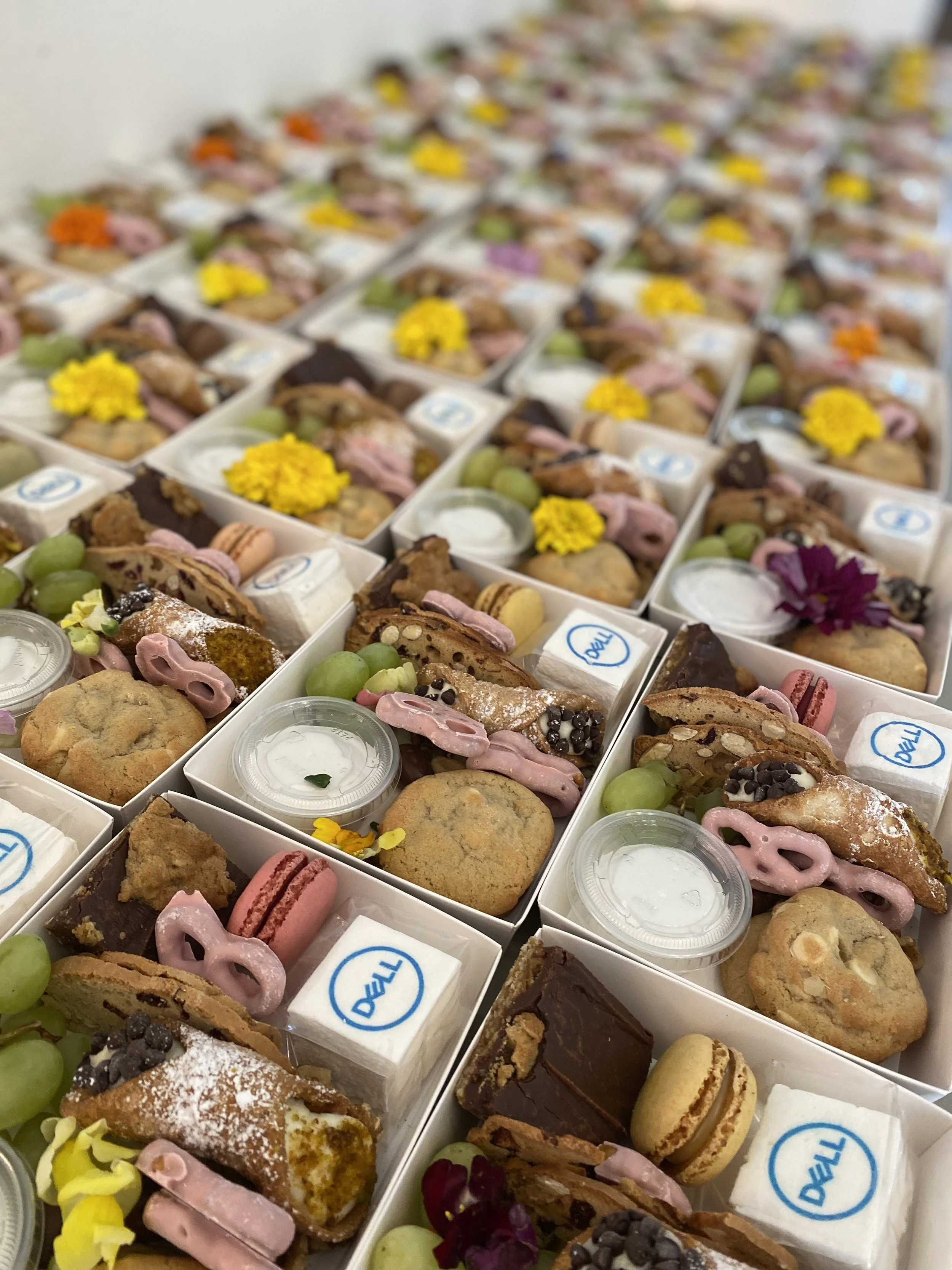 Multiple white boxes filled with assorted cookies, macarons, grapes, and flowers, arranged in a grid pattern.