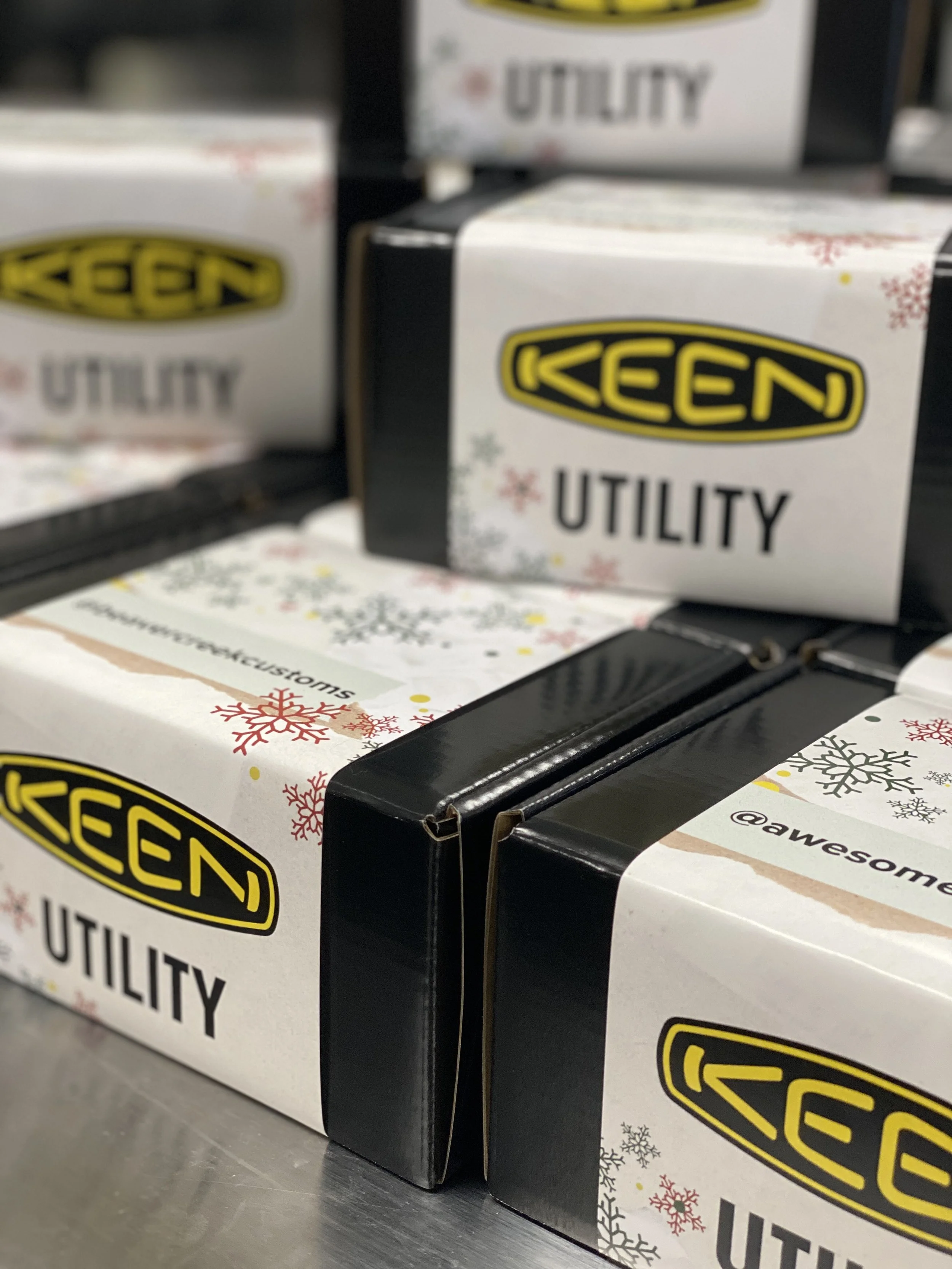 Boxes of KEEN utility shoes with holiday-themed packaging, including snowflakes and the KEEN logo.