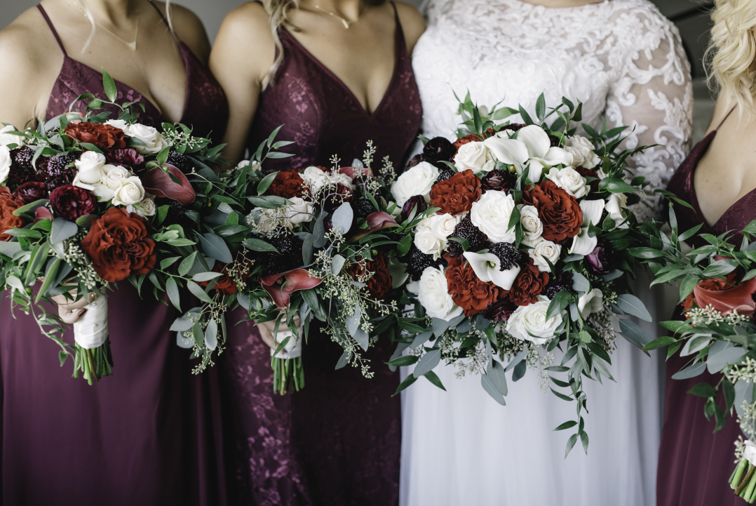Bridal party holding large bouquets of white, red, and dark purple flowers with greenery. Bridesmaids wear burgundy dresses, and bride wears a white lace dress.