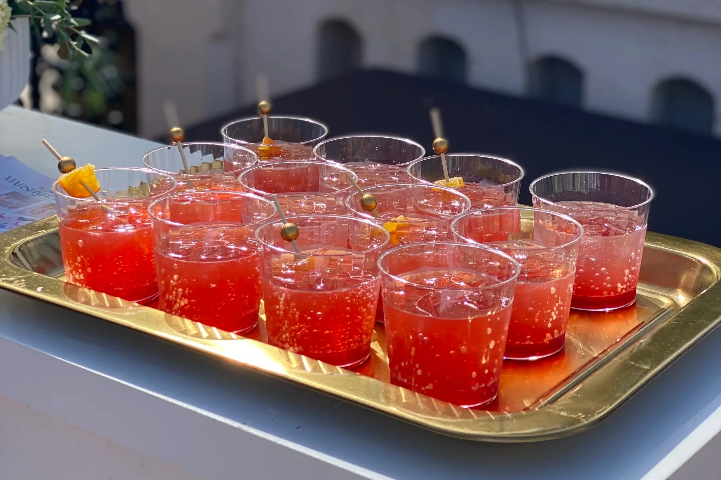 A tray of twelve glasses filled with a red carbonated beverage garnished with pineapple pieces and skewers, set on a white surface outdoors.