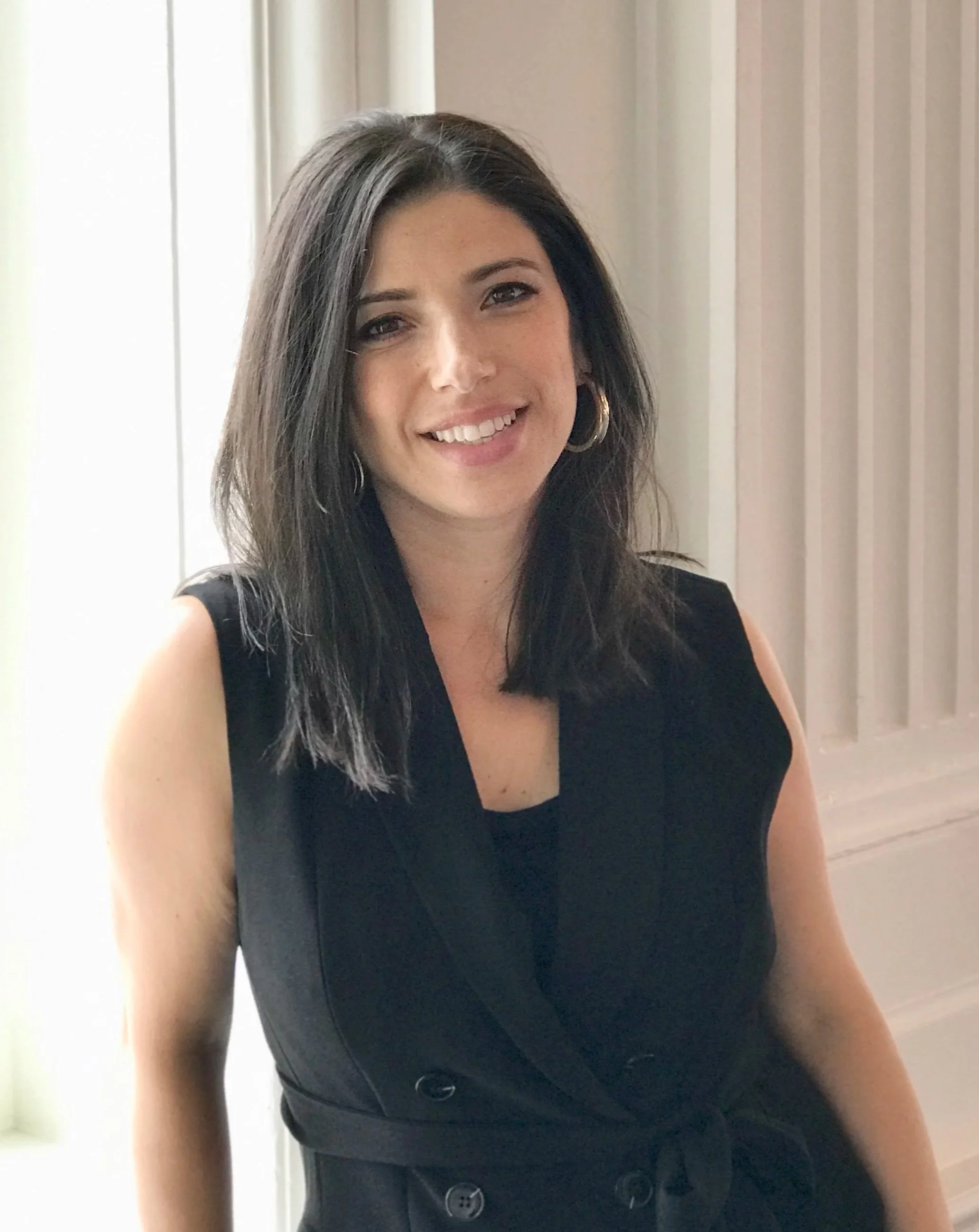 A woman with shoulder-length dark hair smiling at the camera, wearing a sleeveless black dress with a belted waist, gold hoop earrings, standing in front of a window with curtains.