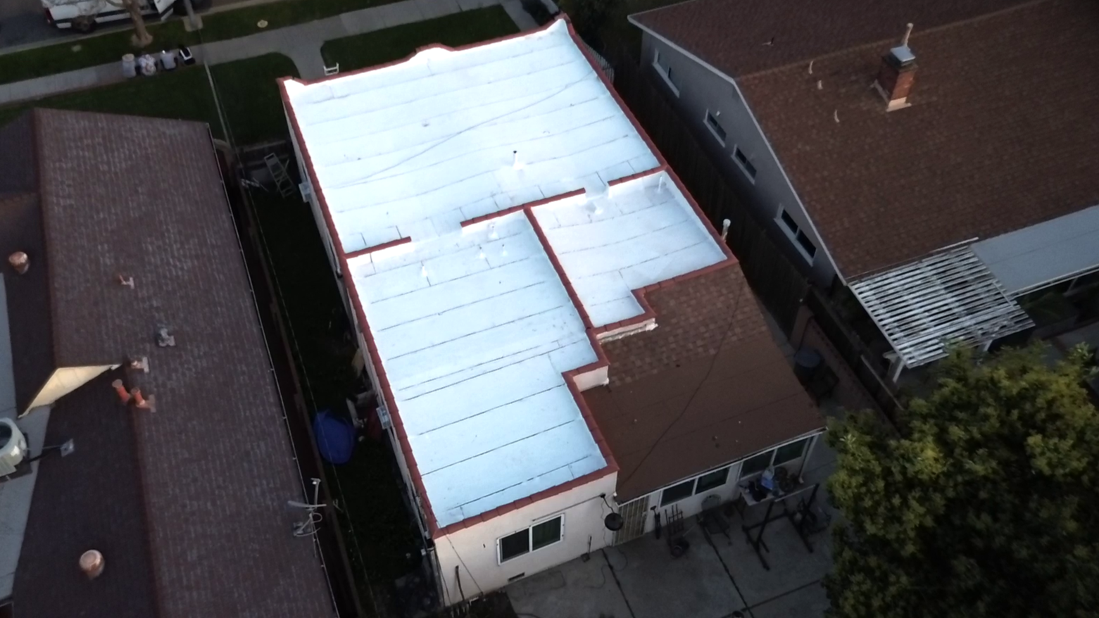 Aerial view of a residential house with a flat white roof, surrounded by neighboring houses and trees.