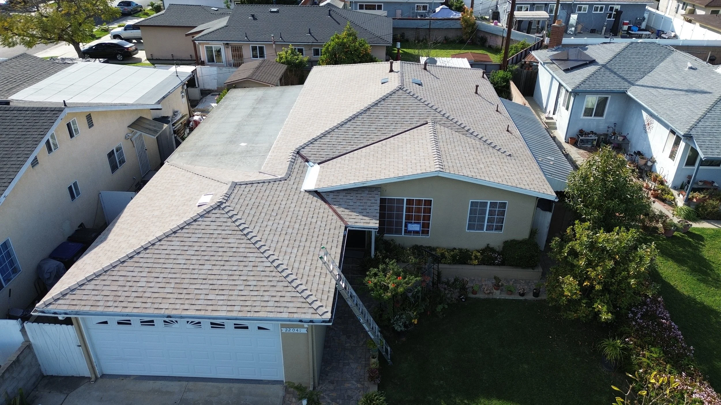 Aerial view of a suburban house with beige and gray roofs, a garage, and a backyard with trees and plants.