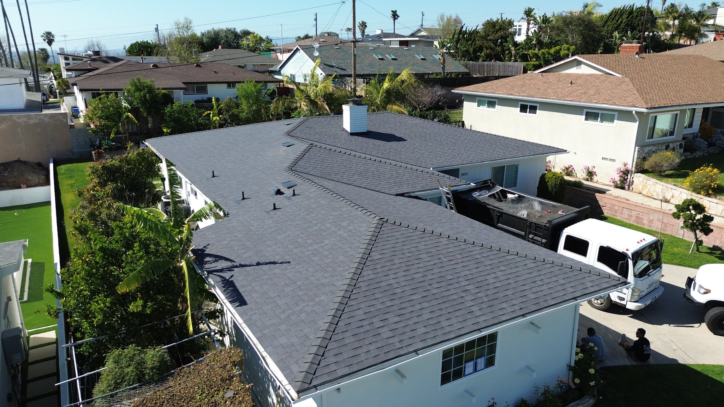 Aerial view of a residential neighborhood featuring a house with a gray shingled roof, surrounded by neighboring houses, trees, and gardens. There is a white truck and a white van parked in the driveway.